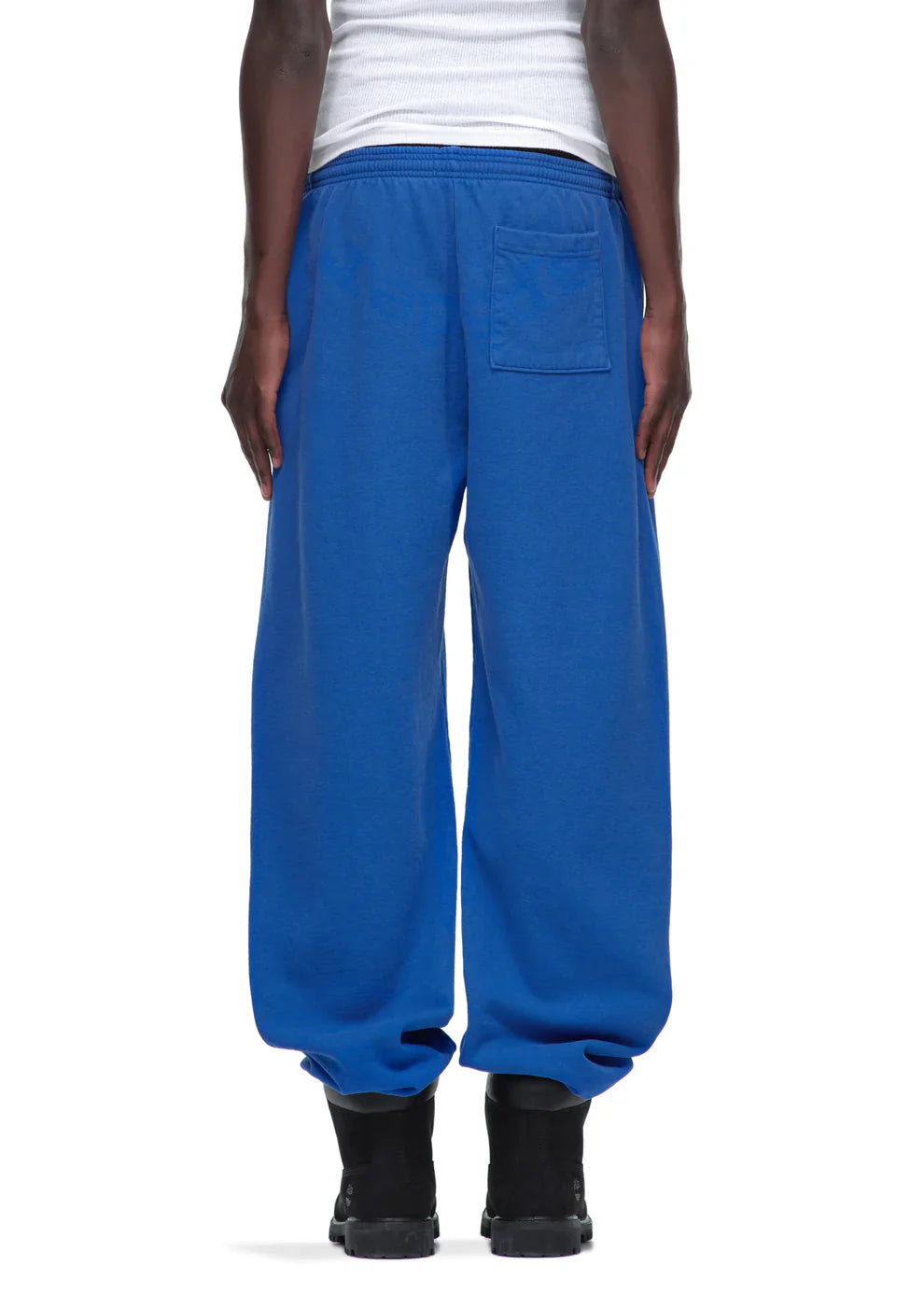 A person wearing loose-fitting Sp5der Sweatpants – Unisex Blue Cotton Sweatpants with a front logo print, paired with a white tank top and black boots, shown from the back for a stylish streetwear look.