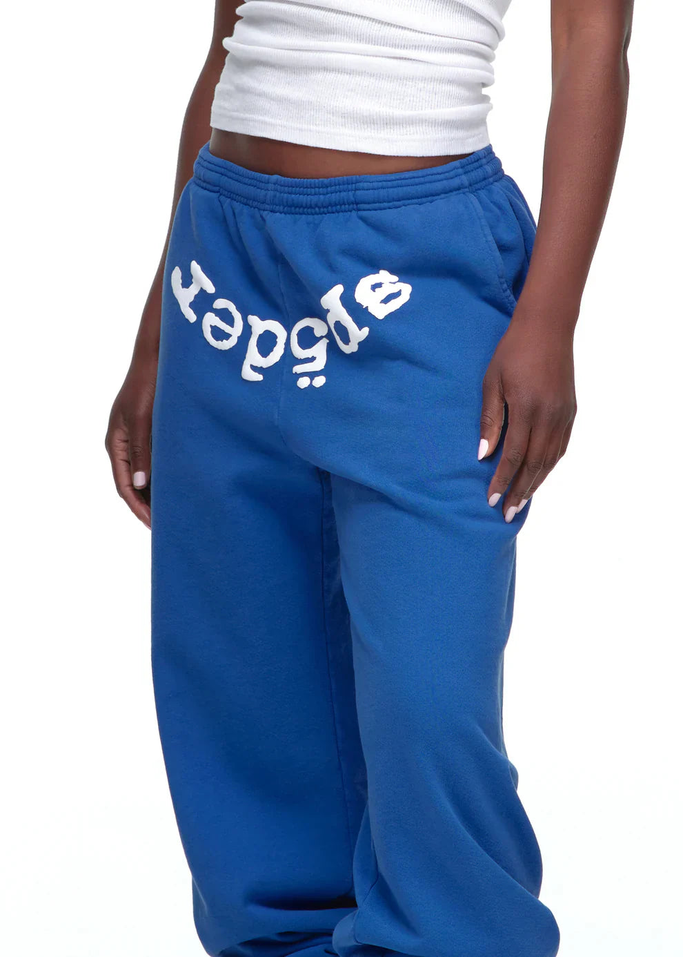 A person wears unisex blue Sp5der Sweatpants with a front logo print and a white ribbed tank top. The streetwear-style image is cropped to show just the lower torso and upper legs.