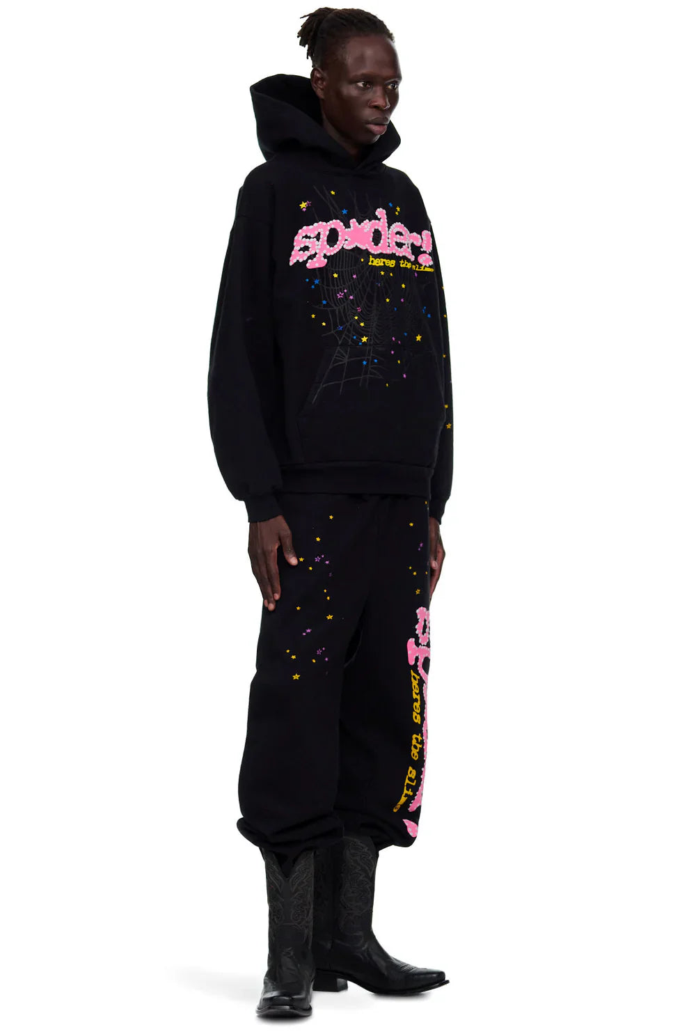 A person stands sideways in a black hoodie and Sp5der Sweatpants—unisex black cotton sweatpants featuring a pink graphic logo and star print—paired with black cowboy boots for a bold streetwear look.