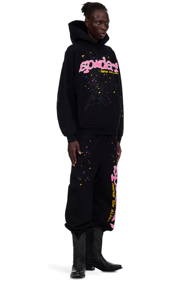 A person stands sideways in a black hoodie and Sp5der Sweatpants—unisex black cotton sweatpants featuring a pink graphic logo and star print—paired with black cowboy boots for a bold streetwear look.