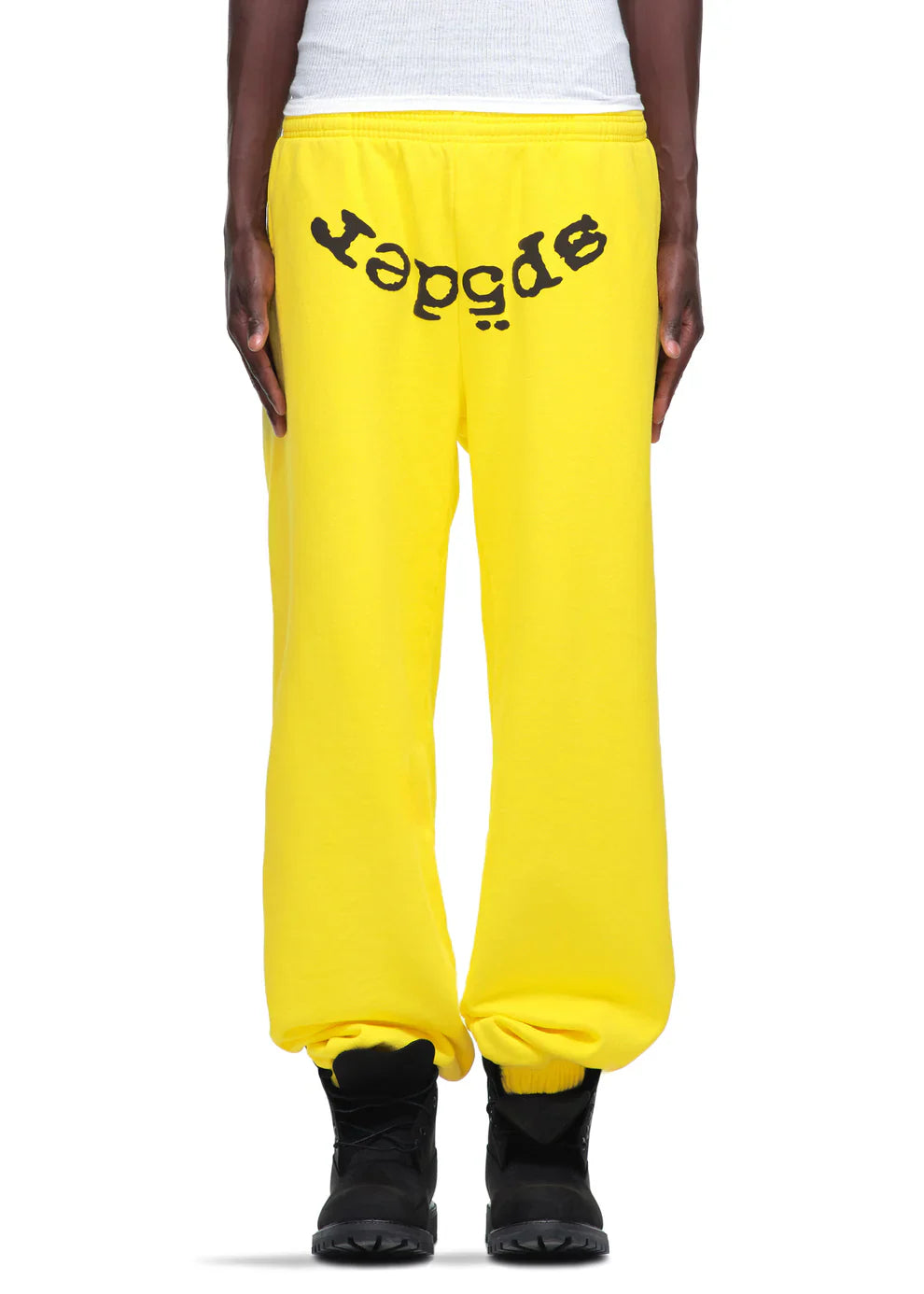 A person wears Sp5der Unisex Yellow Cotton Sweatpants featuring a front logo print, styled as streetwear with black boots, standing with hands relaxed at their sides.