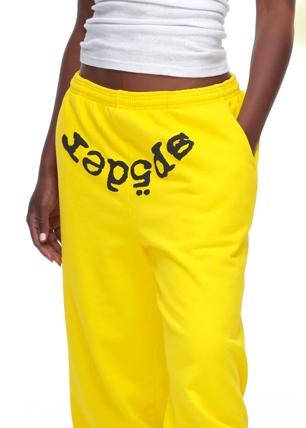 A person sporting Sp5der Sweatpants—unisex yellow cotton sweatpants with a front logo print—and a white ribbed tank top, one hand in their pocket for an effortlessly cool streetwear vibe.