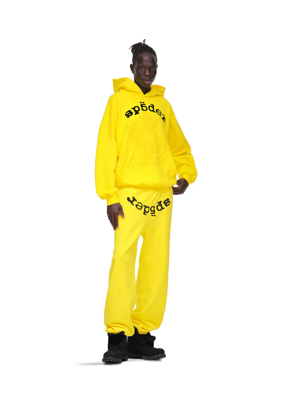 Wearing Sp5der Sweatpants—unisex yellow cotton sweatpants with a front logo print—a person stands against a white background, pairing them with a bright yellow hoodie and black shoes for a bold streetwear look.