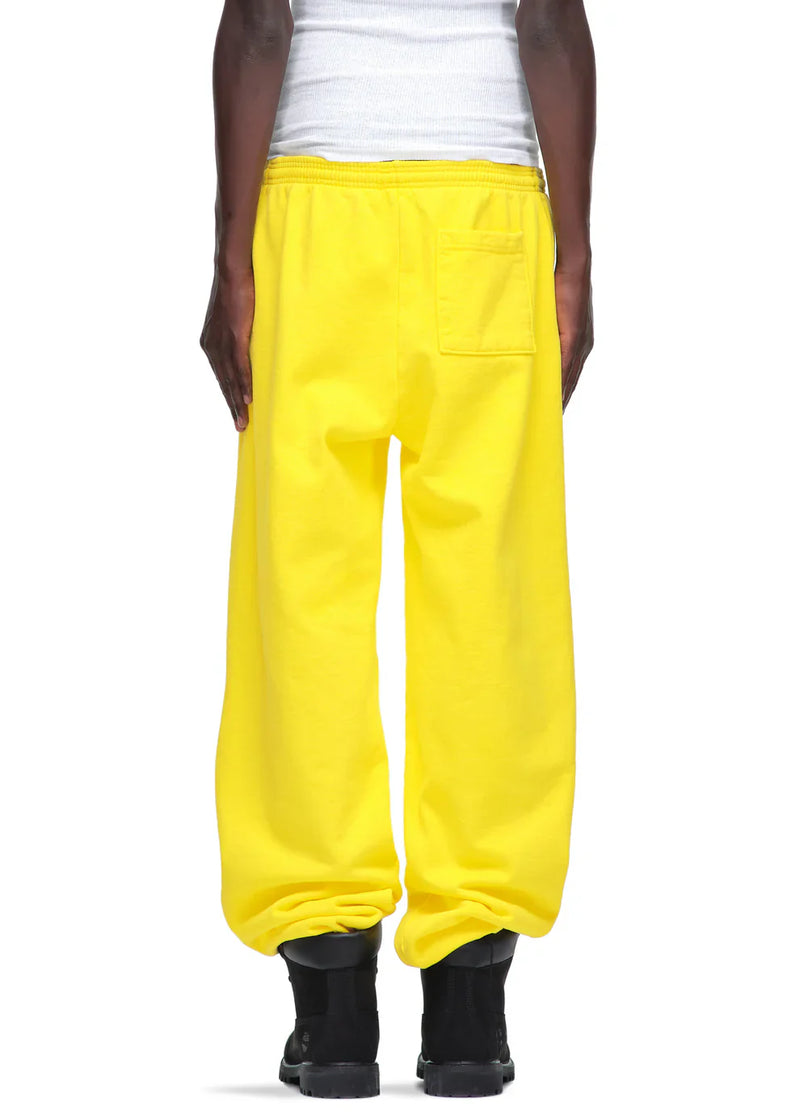 Someone wearing loose Sp5der Sweatpants – Unisex Yellow Cotton Sweatpants with a front logo print, featuring an elastic waistband and back pocket, paired with a white ribbed tank top and black boots for a classic streetwear vibe, shown from the back.
