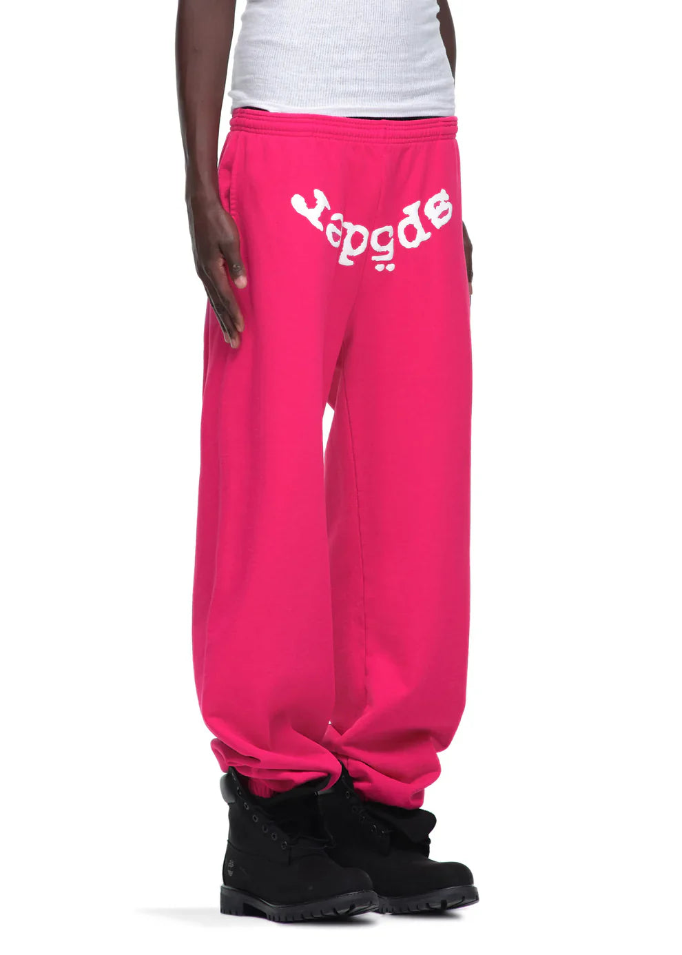 A person styled in Sp5der Sweatpants – Unisex Pink Cotton Sweatpants with a front logo print, paired with a white ribbed tank top and black boots for a striking pink streetwear vibe.