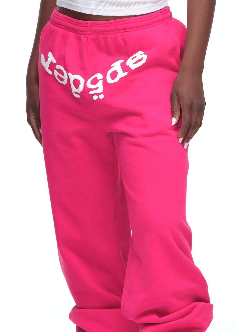A person wears Sp5der Sweatpants – Unisex Pink Cotton Sweatpants with a white front logo print, paired with a white top; only the lower torso and legs are shown, highlighting bold pink streetwear style.