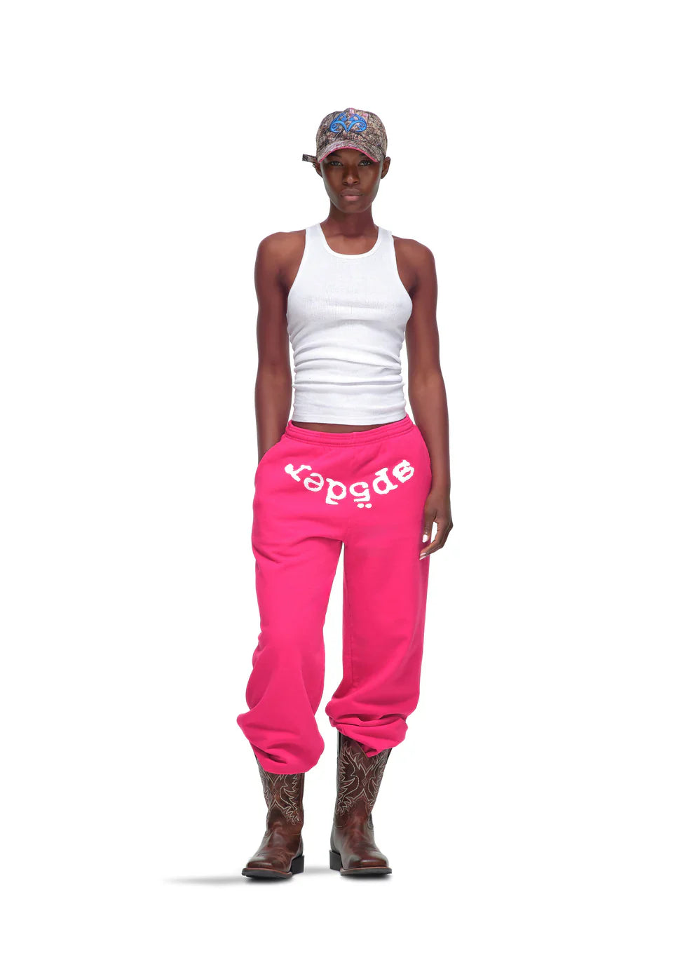 Someone wears a white tank top, Sp5der Sweatpants – Unisex Pink Cotton Sweatpants with Front Logo Print, brown cowboy boots, and a patterned bandana while standing against a plain white background.