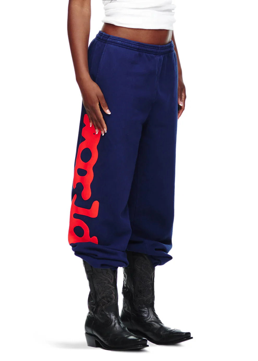 Wearing Sp5der Sweatpants Navy Blue Red Side Logo Print – 100% Cotton Streetwear Jogger Pants, paired with black cowboy boots and a white cropped top, creates a bold streetwear look against a white background.