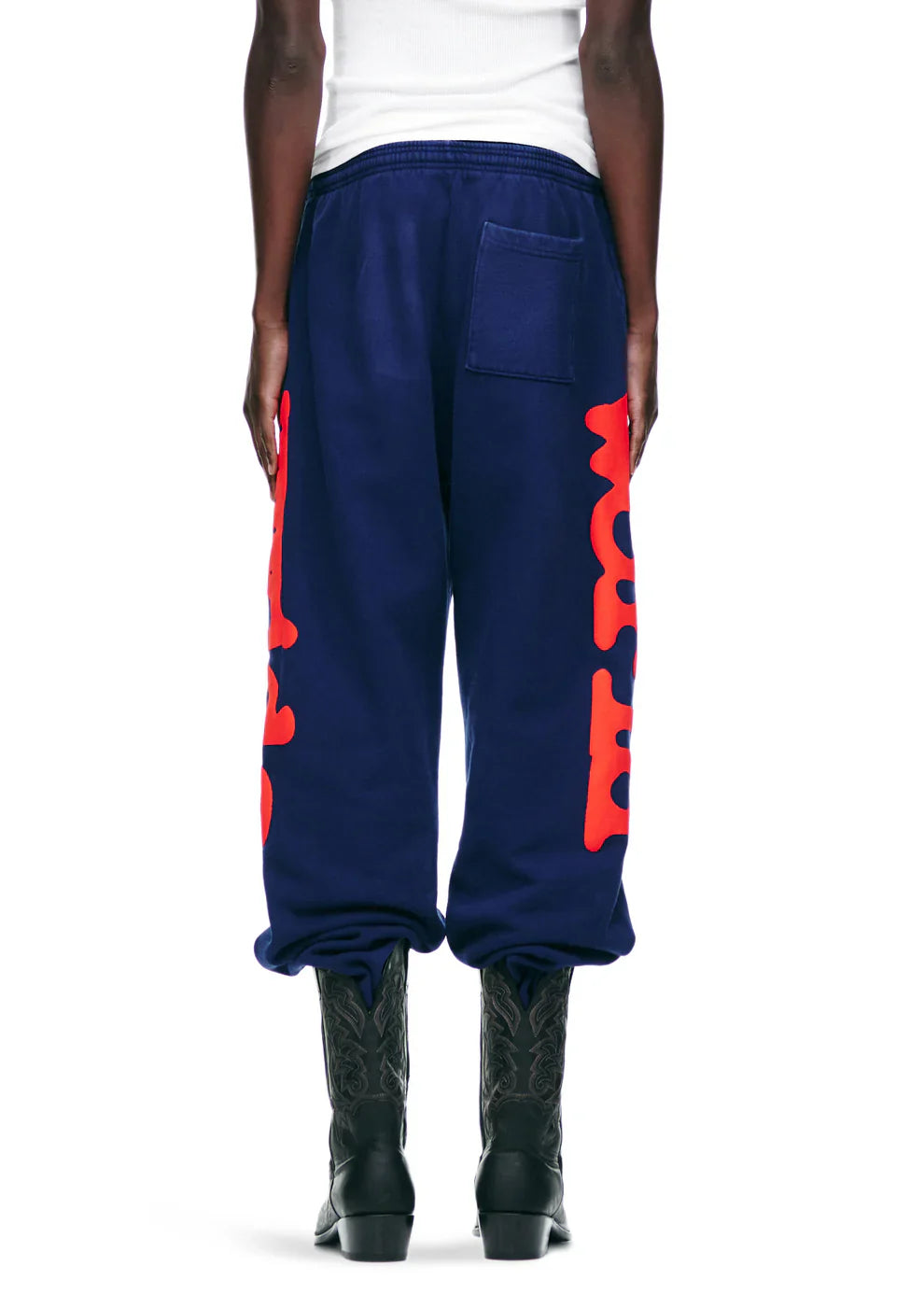Someone wearing Sp5der Sweatpants Navy Blue Red Side Logo Print – 100% Cotton Streetwear Jogger Pants, paired with black cowboy boots and a white top, seen from the back against a plain background for a bold streetwear vibe.