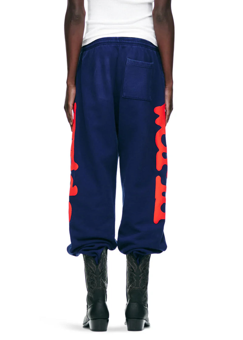 Someone wearing Sp5der Sweatpants Navy Blue Red Side Logo Print – 100% Cotton Streetwear Jogger Pants, paired with black cowboy boots and a white top, seen from the back against a plain background for a bold streetwear vibe.