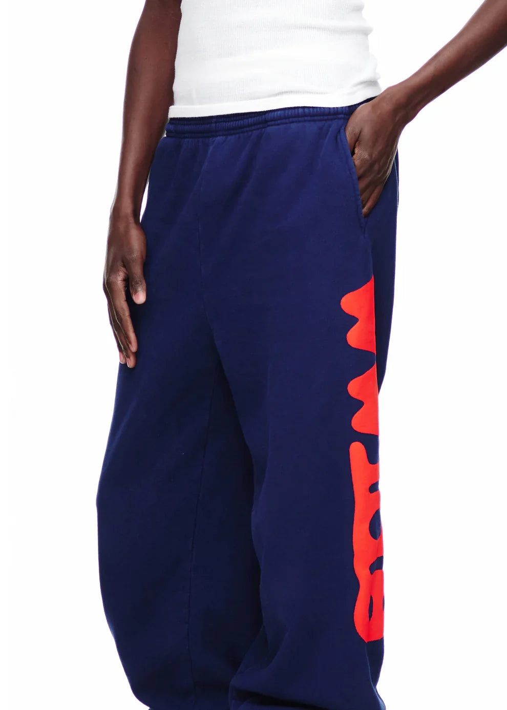 A person wearing Sp5der Sweatpants Navy Blue Red Side Logo Print—100% cotton jogger pants—with a bold red graphic, paired with a white sleeveless shirt, stands in streetwear style with one hand in their pocket.