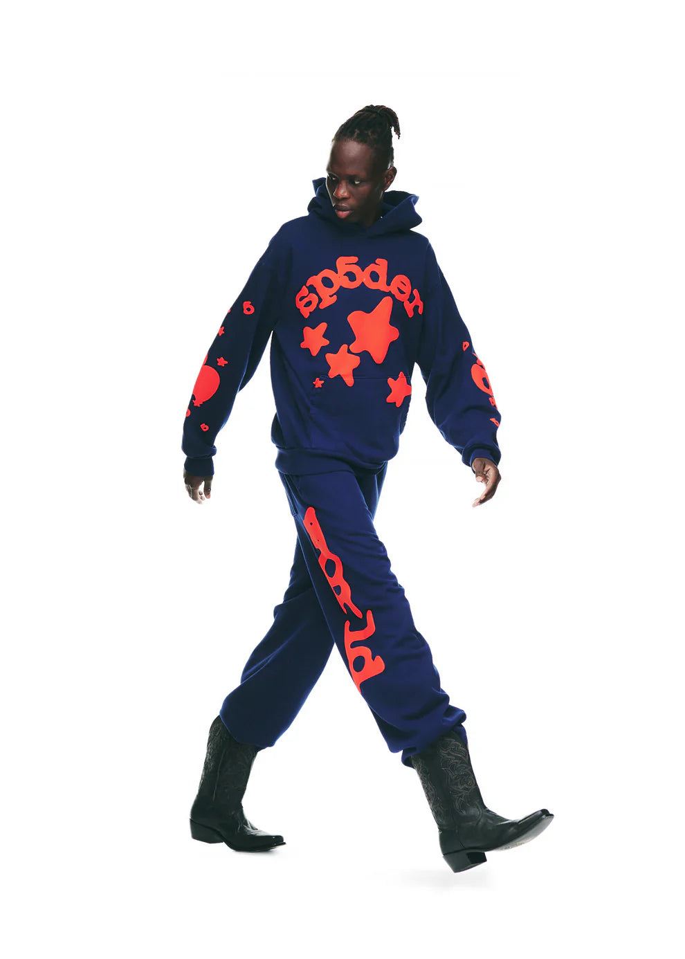 Wearing a navy blue hoodie and Sp5der Sweatpants Navy Blue Red Side Logo Print – 100% Cotton Streetwear Jogger Pants, paired with black cowboy boots, a person walks confidently against a white background, channeling standout streetwear energy.