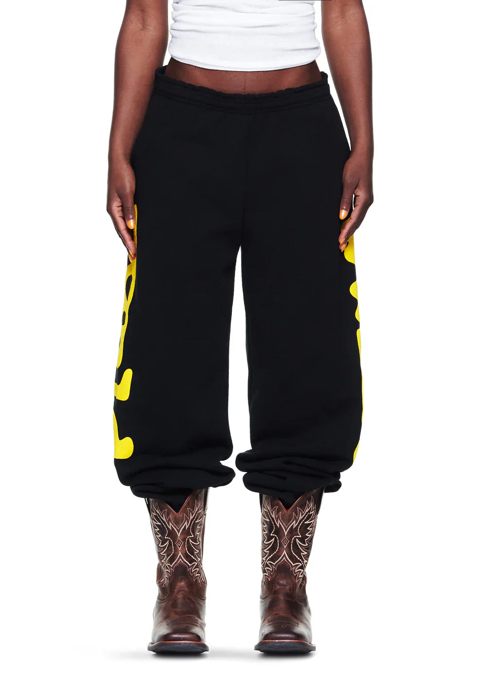 A person wearing a white shirt, Sp5der Sweatpants Black Yellow Side Logo Print – 100% Cotton Streetwear Jogger Pants, and brown embroidered cowboy boots stands against a white background.