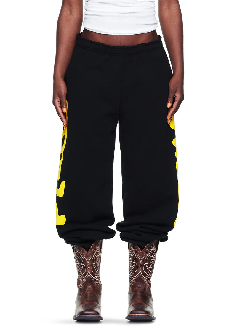 A person wearing a white shirt, Sp5der Sweatpants Black Yellow Side Logo Print – 100% Cotton Streetwear Jogger Pants, and brown embroidered cowboy boots stands against a white background.