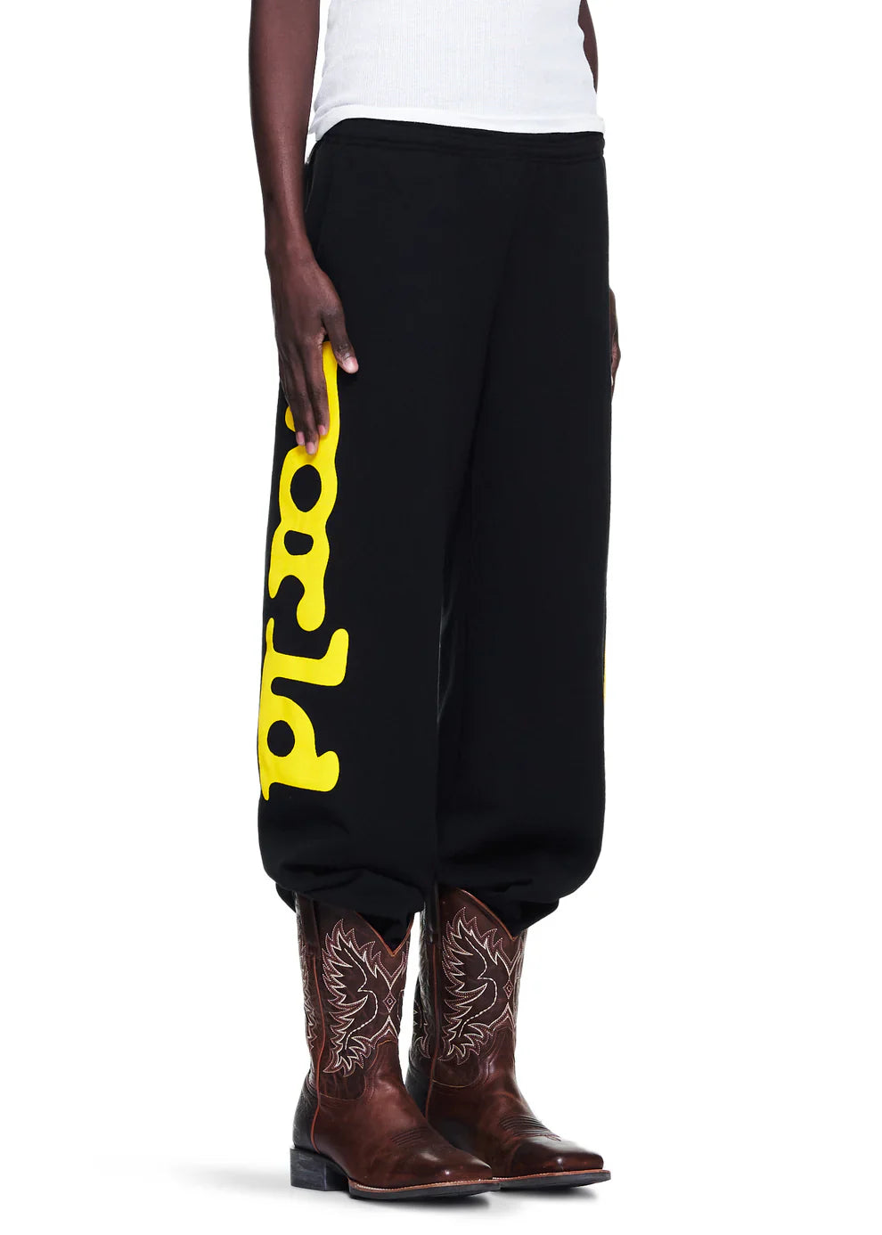 A person sports Sp5der Sweatpants Black Yellow Side Logo Print (100% cotton streetwear jogger pants) with brown cowboy boots and a white sleeveless shirt—the upper body is partially visible, adding a bold twist to this street look.