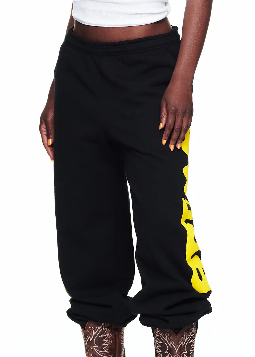 Wearing Sp5der Sweatpants Black Yellow Side Logo Print—100% cotton streetwear jogger pants—a person pairs them with a white crop top and brown patterned boots, standing against a white background in casual style.