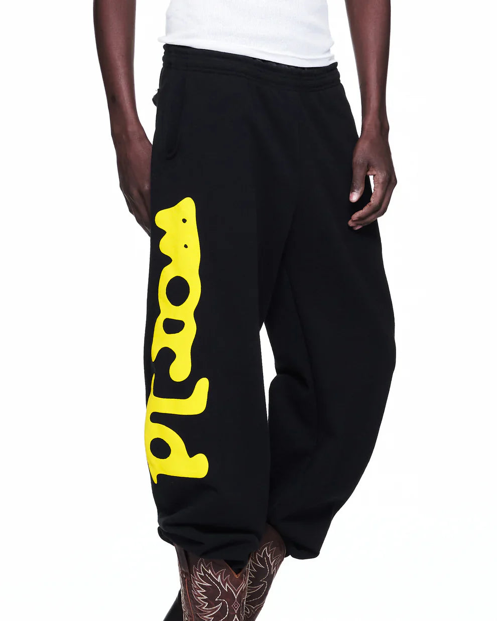 A person wears Sp5der Sweatpants Black Yellow Side Logo Print – 100% Cotton Streetwear Jogger Pants with a white shirt. Tattooed lower legs enhance the edgy vibe of these bold black joggers featuring yellow graphic text along one leg.