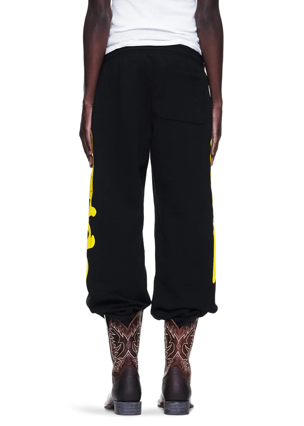 A person stands with their back turned, wearing Sp5der Sweatpants Black Yellow Side Logo Print—100% cotton streetwear jogger pants—rolled at the ankles, paired with brown patterned cowboy boots and a white shirt for a bold street-western look.