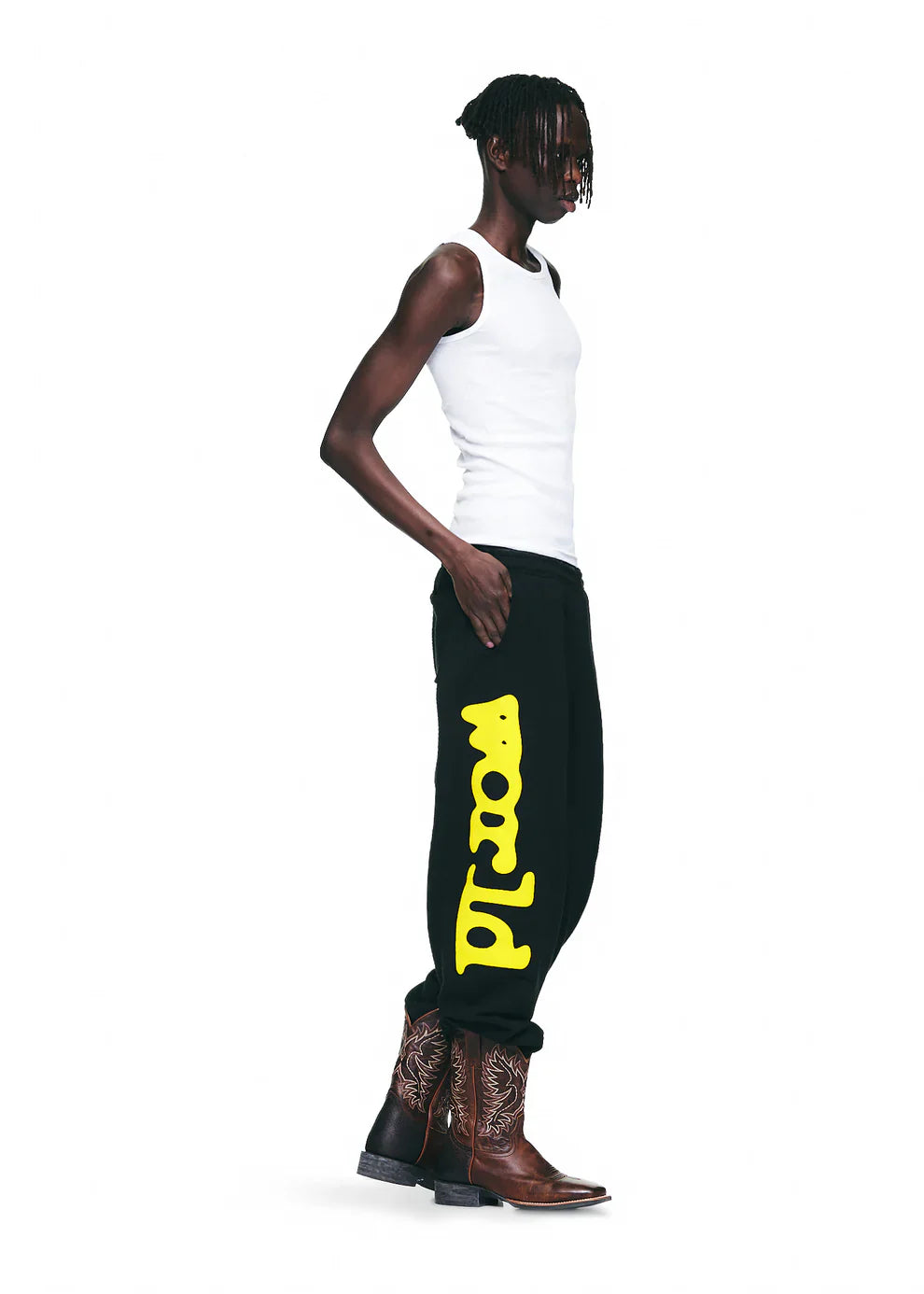 A person wears a white tank top, Sp5der Sweatpants Black Yellow Side Logo Print – 100% Cotton Streetwear Jogger Pants, and brown cowboy boots, standing against a plain white background.