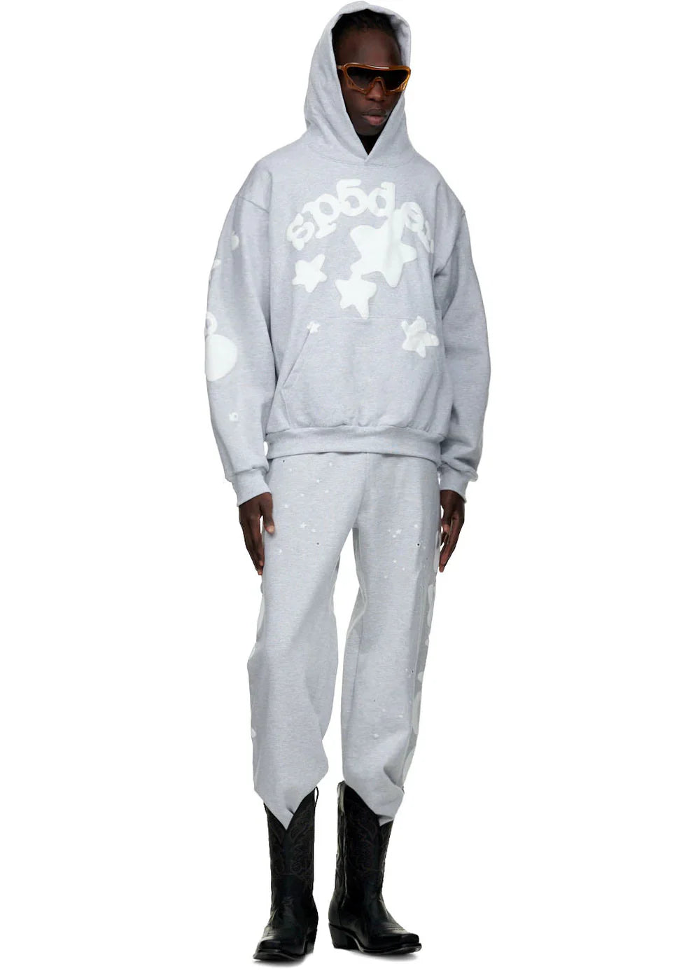 A person stands against a white background wearing orange sunglasses, a light gray hoodie with white graphics, black cowboy boots, and 100% Cotton Sp5der Sweatpants for Men – Grey Streetwear Jogger Pants with Graphic Print.