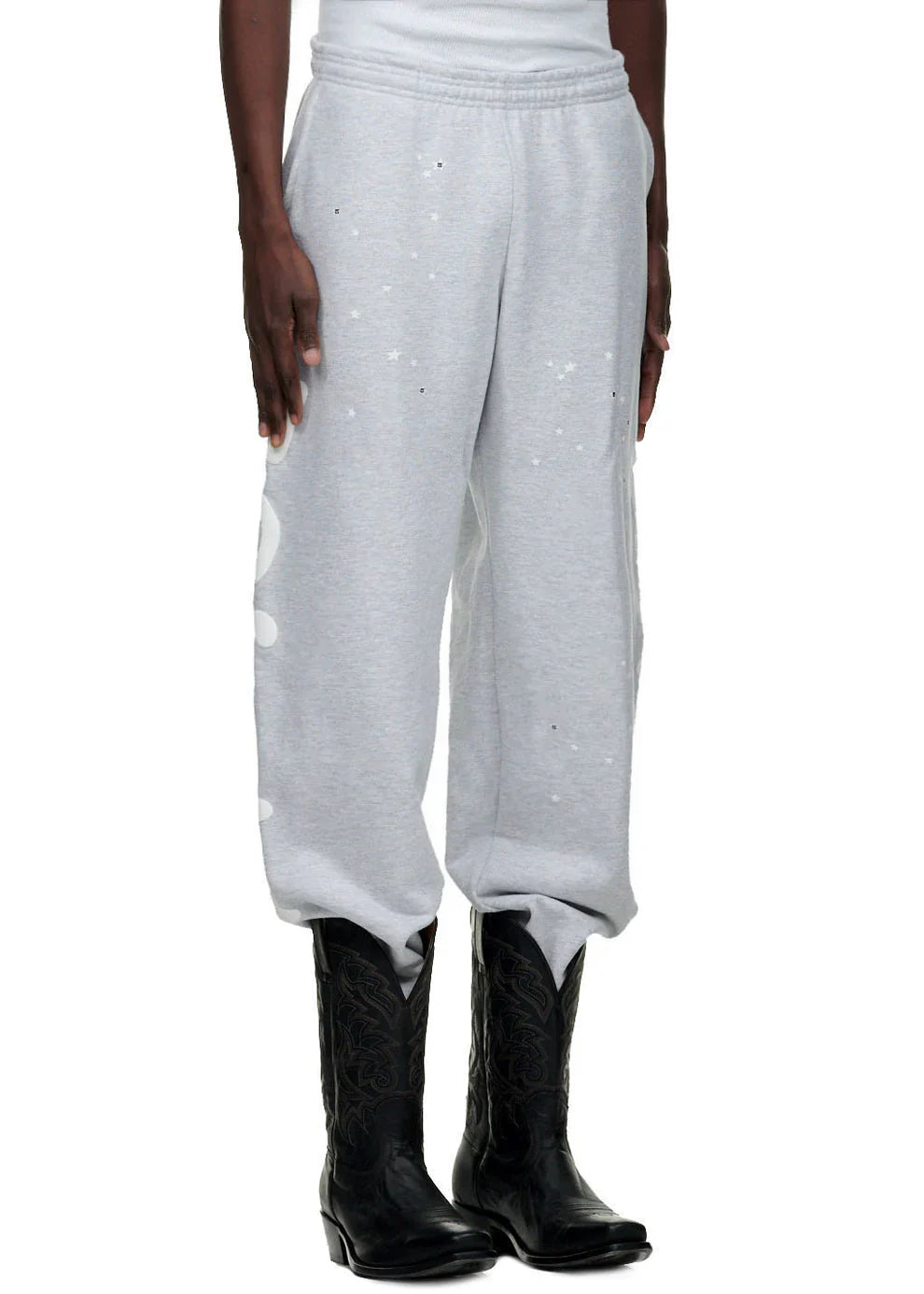 A person wearing 100% Cotton Sp5der Sweatpants for Men – Grey Streetwear Jogger Pants with Graphic Print, paired with a white top and black cowboy boots, showcases a casual streetwear look against a white background.