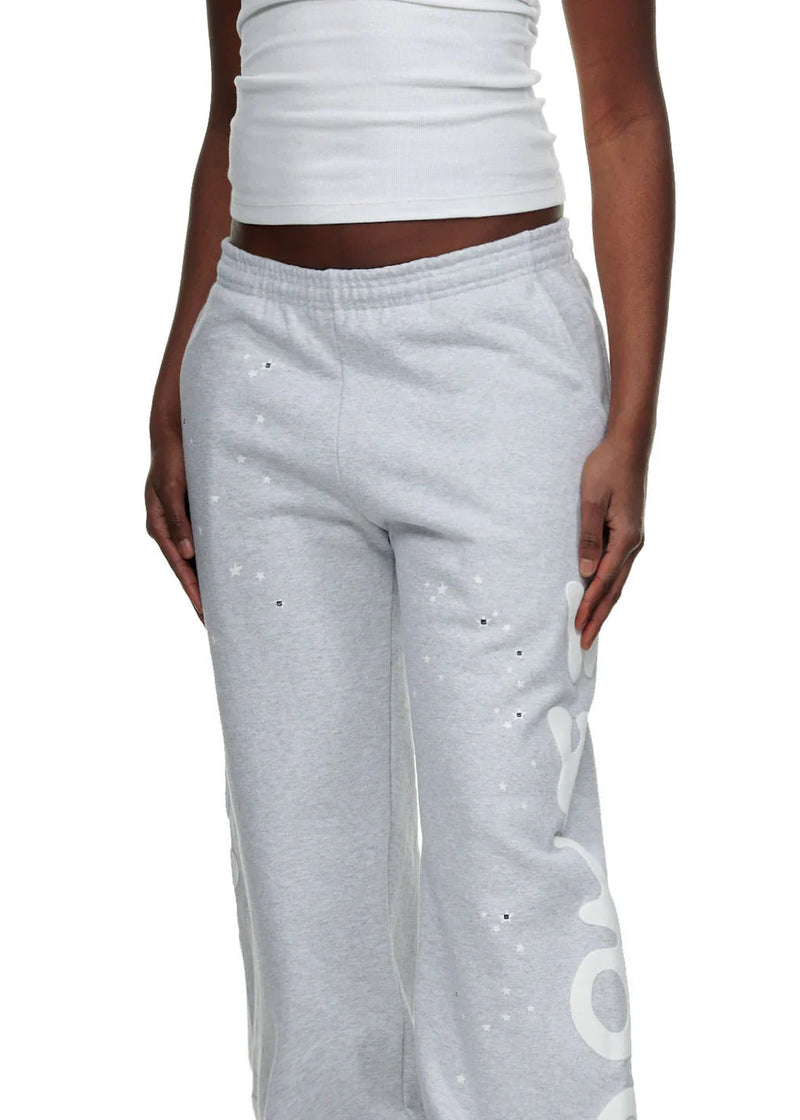 A person stands against a plain background wearing a white ribbed tank top and 100% Cotton Sp5der Sweatpants for Men—Grey Streetwear Jogger Pants with Graphic Print and relaxed fit, showcasing laid-back streetwear style.