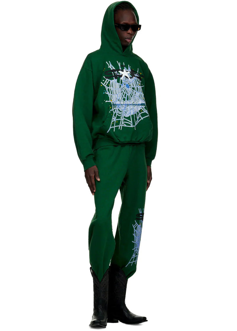 A person stands against a white background wearing black sunglasses, black cowboy boots, and 100% Cotton Sp5der Sweatpants for Men—Dark Green Streetwear Jogger Pants with Spider Web Graphic, Relaxed Fit.
