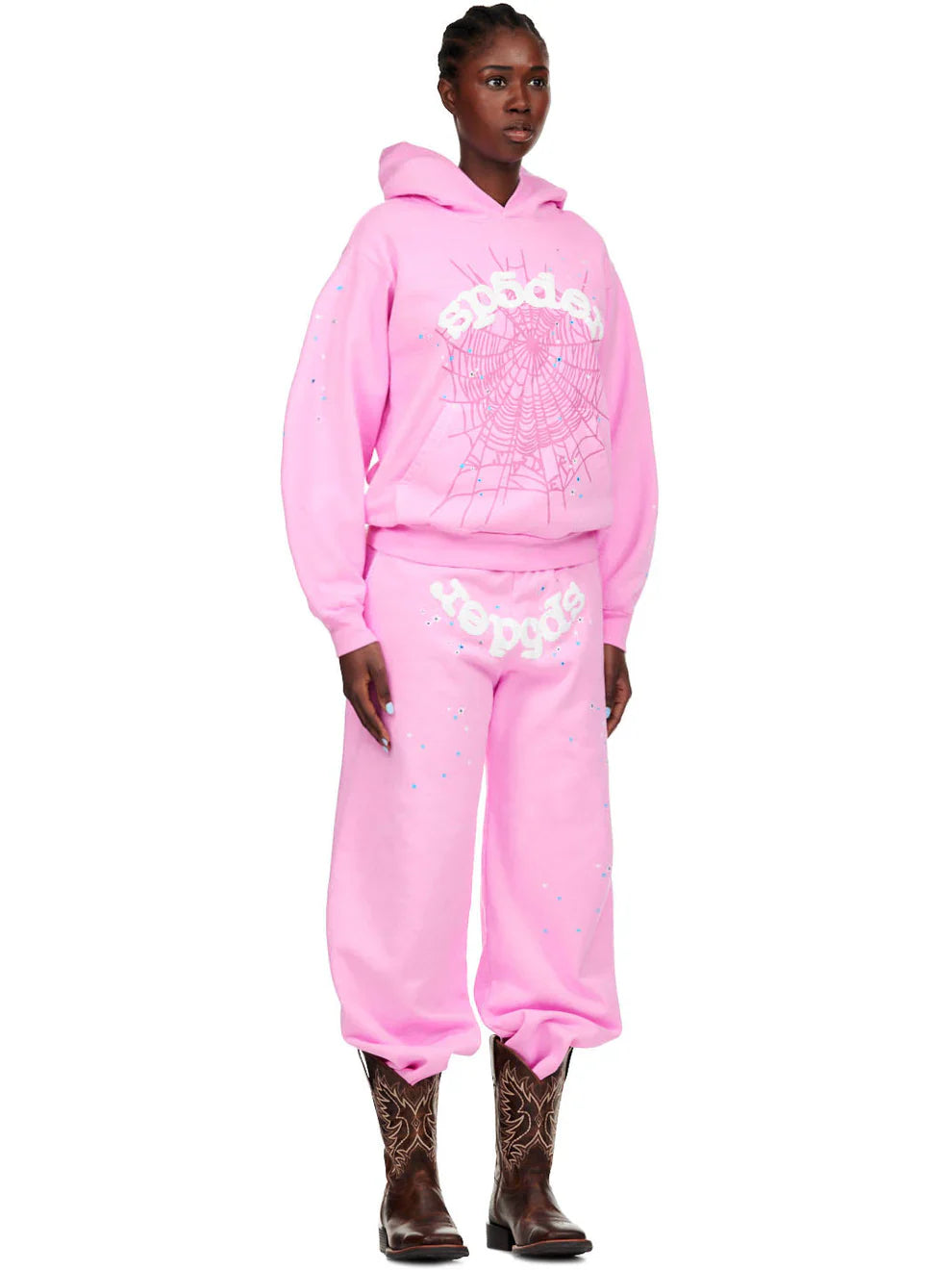 A person stands in bold streetwear—a loose-fitting, unisex jogger hoodie set with white graphics and blue accents, brown cowboy boots, and Sp5der Sweatpants: light pink unisex cotton sweatpants with front logo and star print.