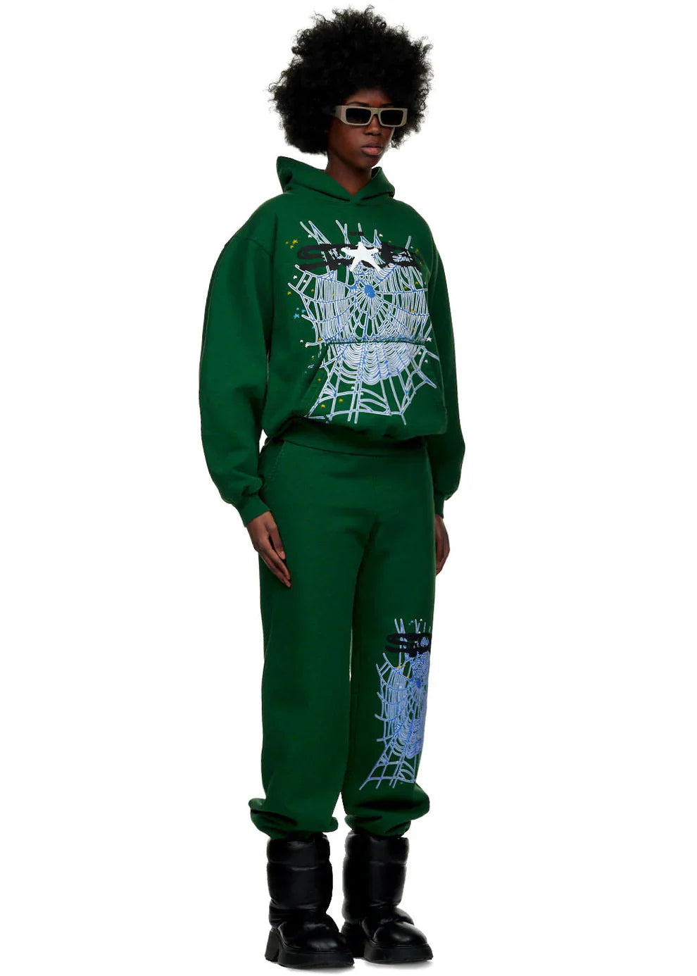 A person in 100% Cotton Sp5der Sweatpants for Men—dark green streetwear jogger pants with spider web graphics and relaxed fit—stands facing forward, styled with a matching hoodie, black boots, large sunglasses, and an afro.