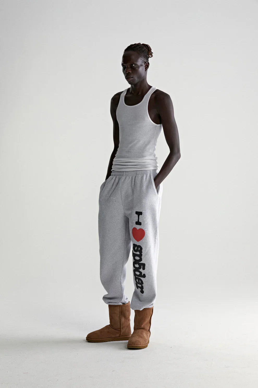 A person with dark skin wears a light gray tank top, 100% Cotton Sp5der Sweatpants for Men—light grey jogger pants with an “I Love Sp5der” graphic, relaxed fit—and brown boots, standing against a plain, light background with hands in pockets.