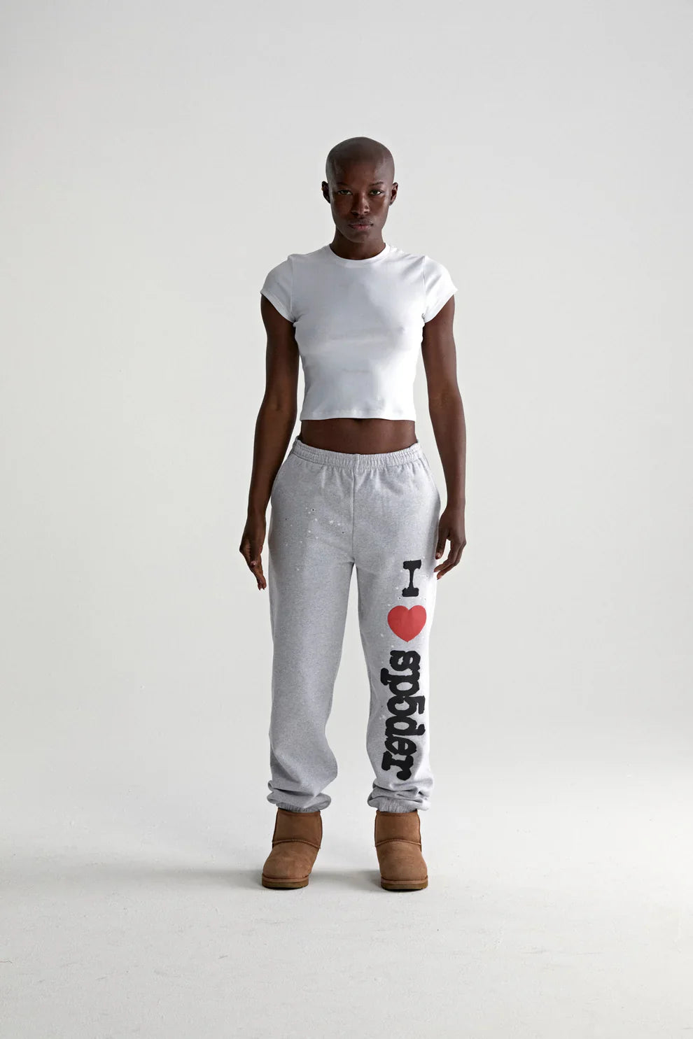 A person faces forward in a fitted white crop top, brown boots, and light grey Sp5der jogger pants featuring “I Love Sp5der” graphic, relaxed fit—showcasing casual streetwear style against a plain white backdrop.