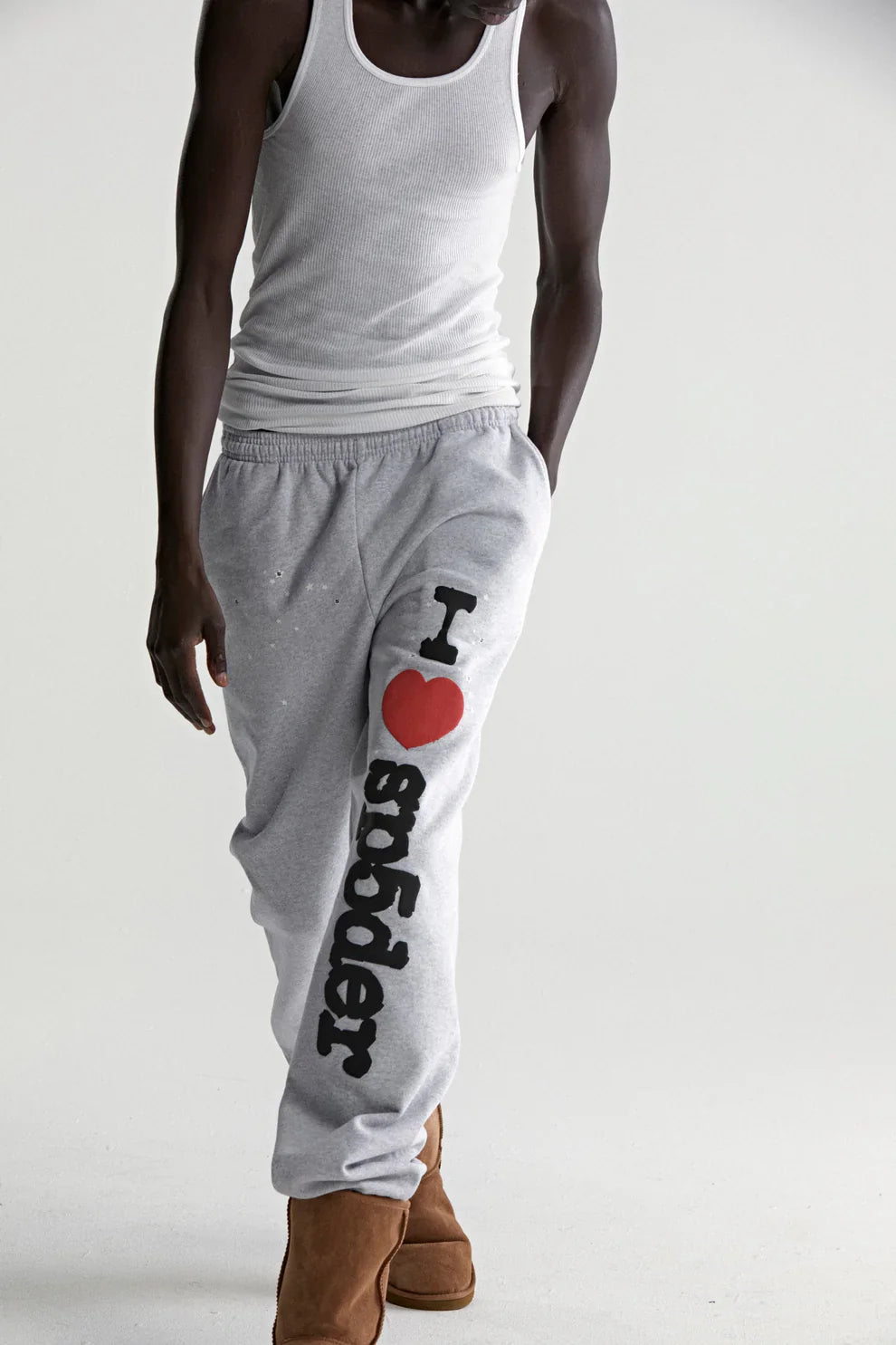 A person wearing a white tank top, brown boots, and 100% cotton Sp5der sweatpants for men in light grey with an "I Love Sp5der" graphic stands against a plain light background, showcasing relaxed streetwear style.