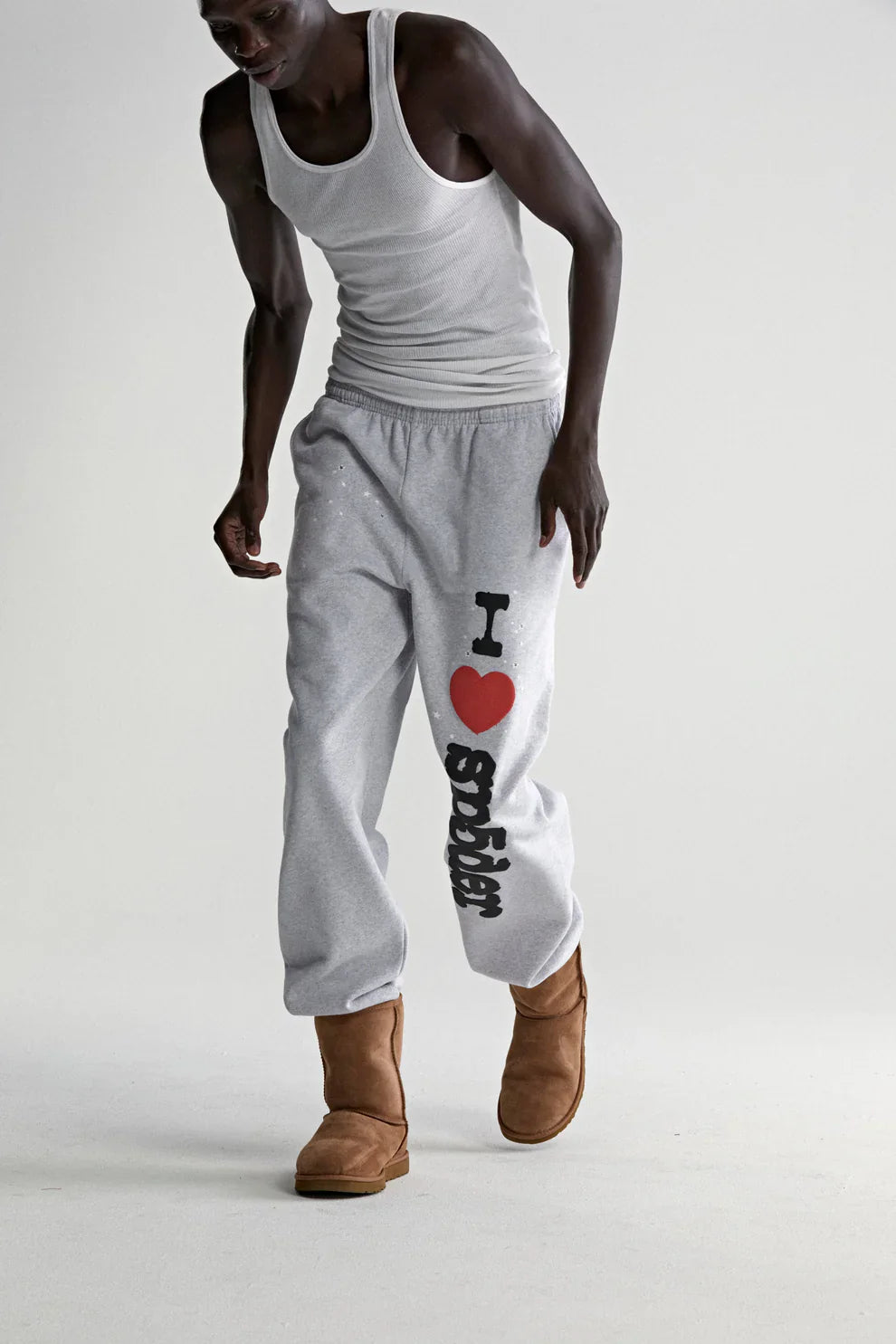 A person in a white tank top, tan UGG-style boots, and 100% Cotton Sp5der Sweatpants for Men—light grey joggers with “I Love Sp5der” graphic—stands against a plain light background, highlighting laid-back streetwear style.