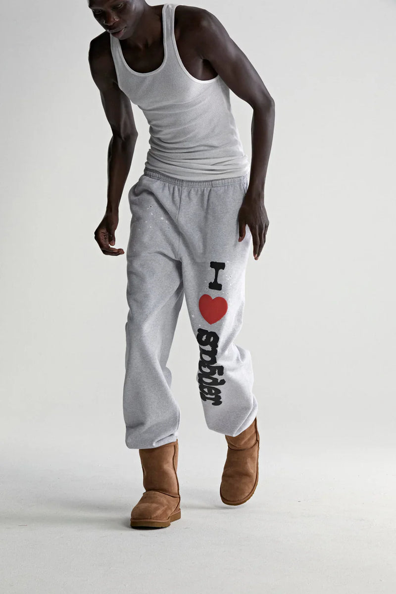 A person in a white tank top, tan UGG-style boots, and 100% Cotton Sp5der Sweatpants for Men—light grey joggers with “I Love Sp5der” graphic—stands against a plain light background, highlighting laid-back streetwear style.