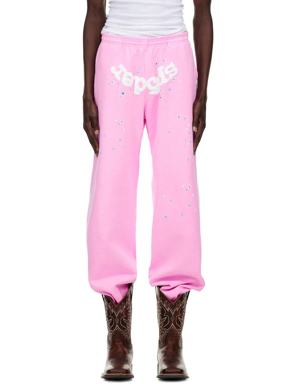 A person wears Sp5der Sweatpants – Unisex Light Pink Cotton Sweatpants with front logo and star print, paired with a white tank top and brown cowboy boots, standing against a white background.