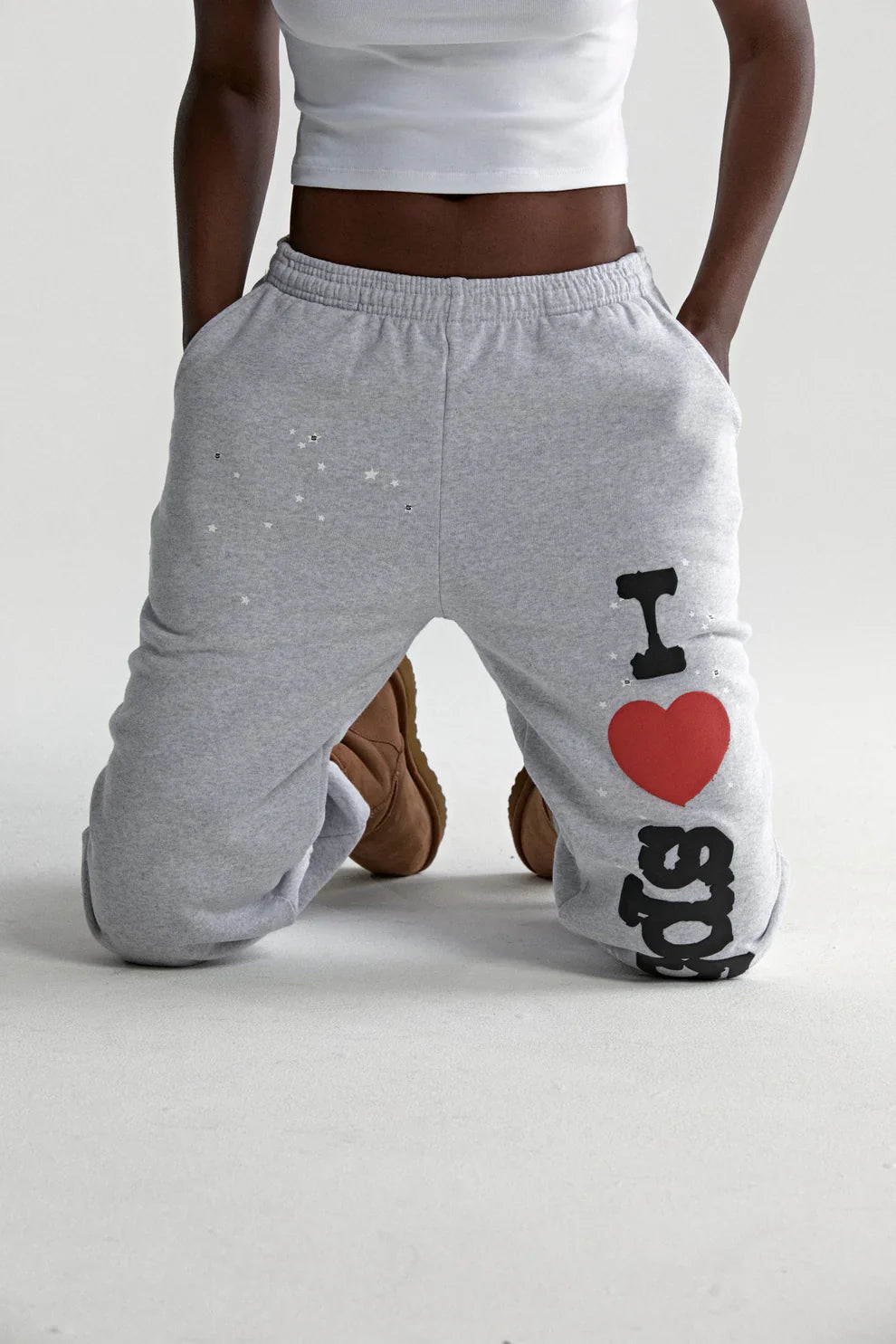 Someone kneels on the floor in 100% Cotton Sp5der Sweatpants for Men—light grey joggers with an “I Love Sp5der” graphic and relaxed fit—paired with a white cropped top and brown boots for a trendy streetwear look.