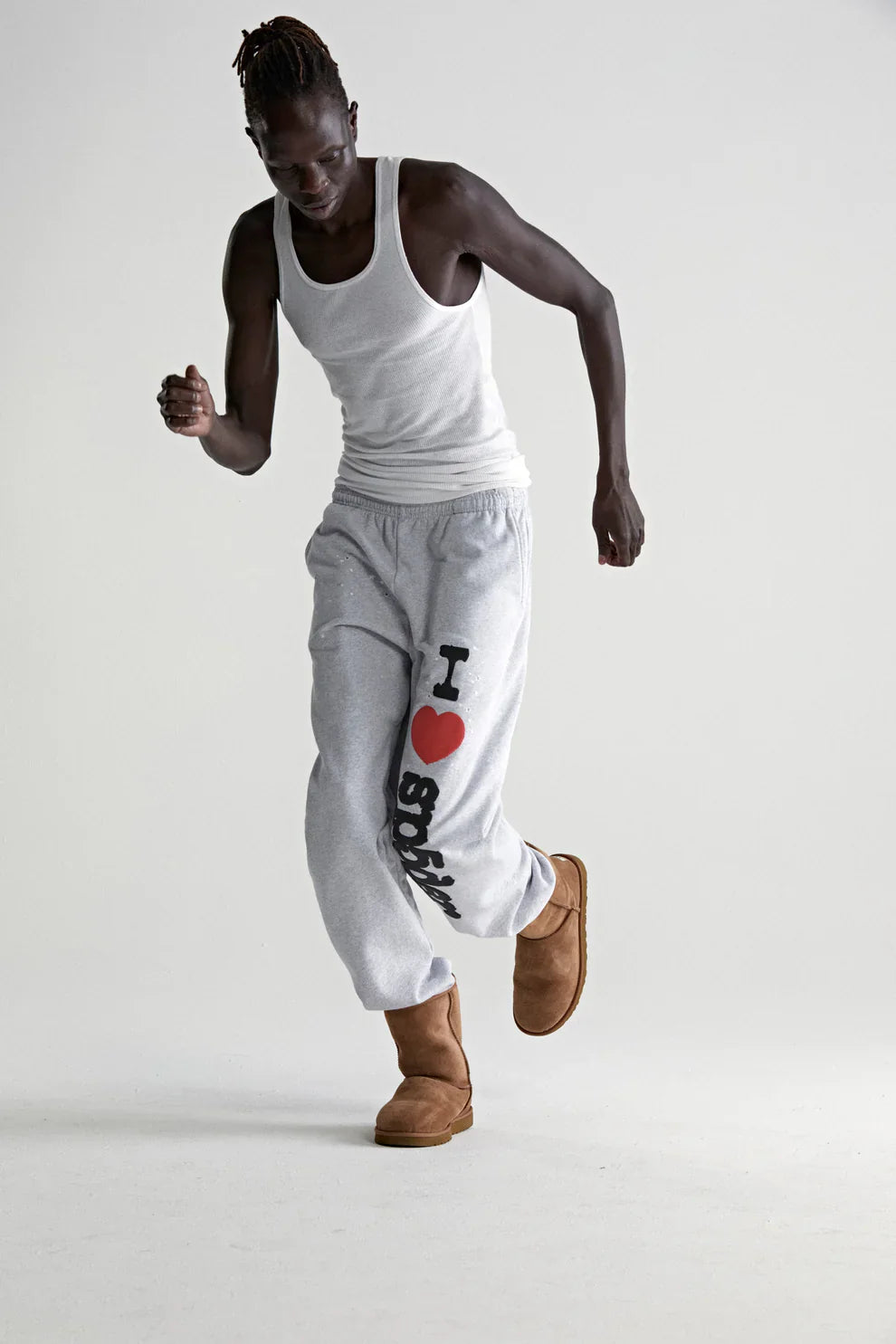 A person in a white tank top, 100% Cotton Sp5der Sweatpants for Men—light grey joggers with an “I Love Sp5der” graphic and relaxed fit—and brown slippers poses against a plain light background, showcasing effortless streetwear style.