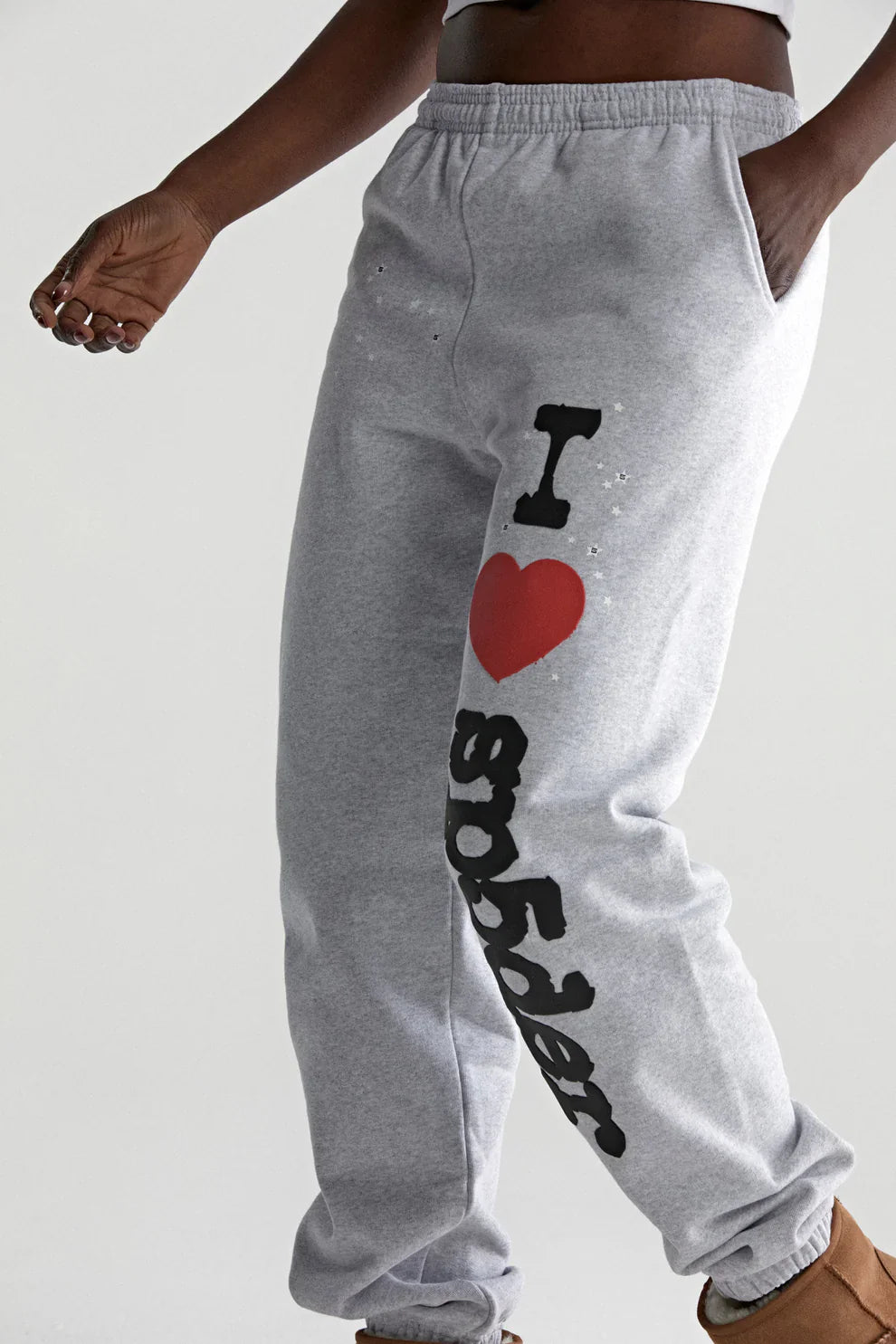 A man sports light grey 100% cotton Sp5der Sweatpants, featuring a relaxed fit and "I Love Sp5der" graphic on one leg. With one hand in his pocket and brown shoes, he showcases an easygoing streetwear style.
