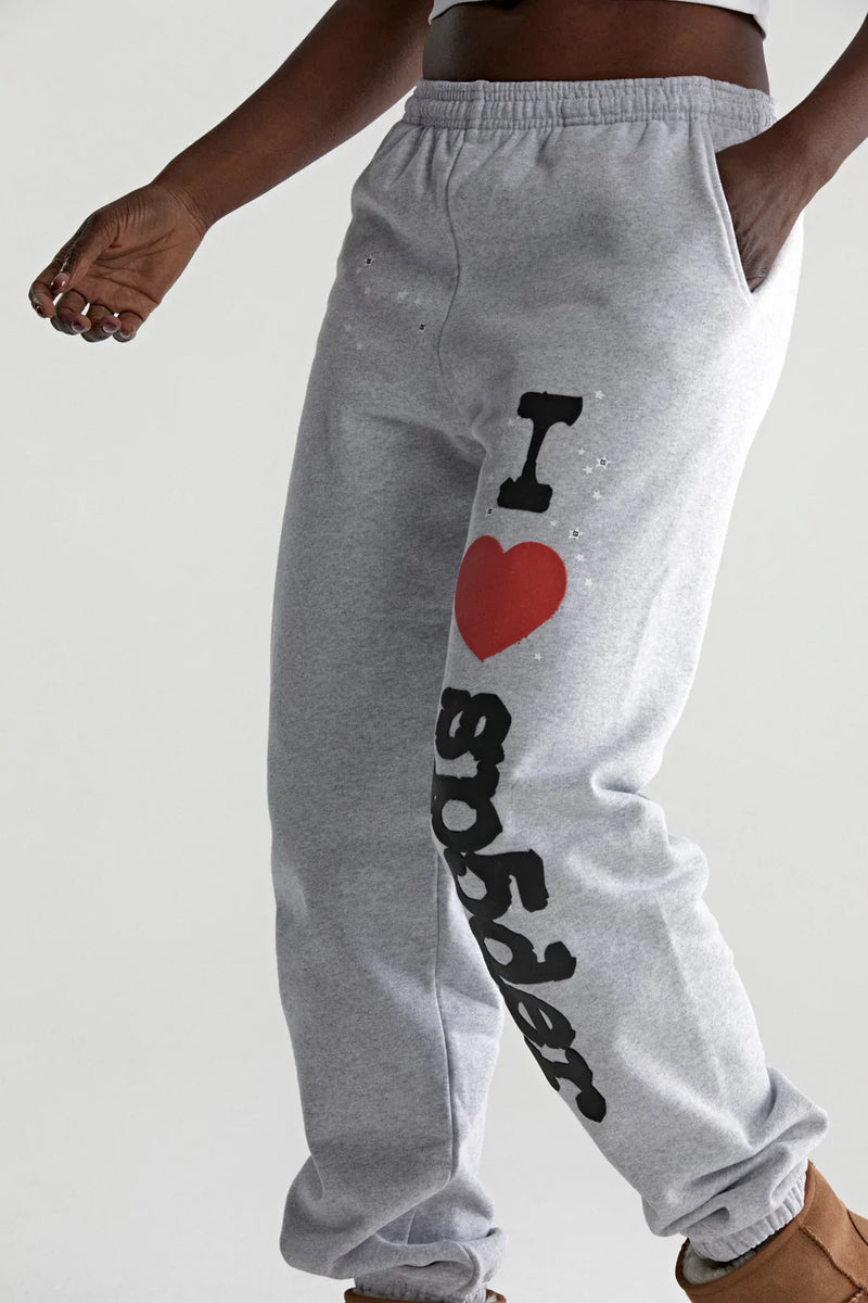 A man sports light grey 100% cotton Sp5der Sweatpants, featuring a relaxed fit and "I Love Sp5der" graphic on one leg. With one hand in his pocket and brown shoes, he showcases an easygoing streetwear style.