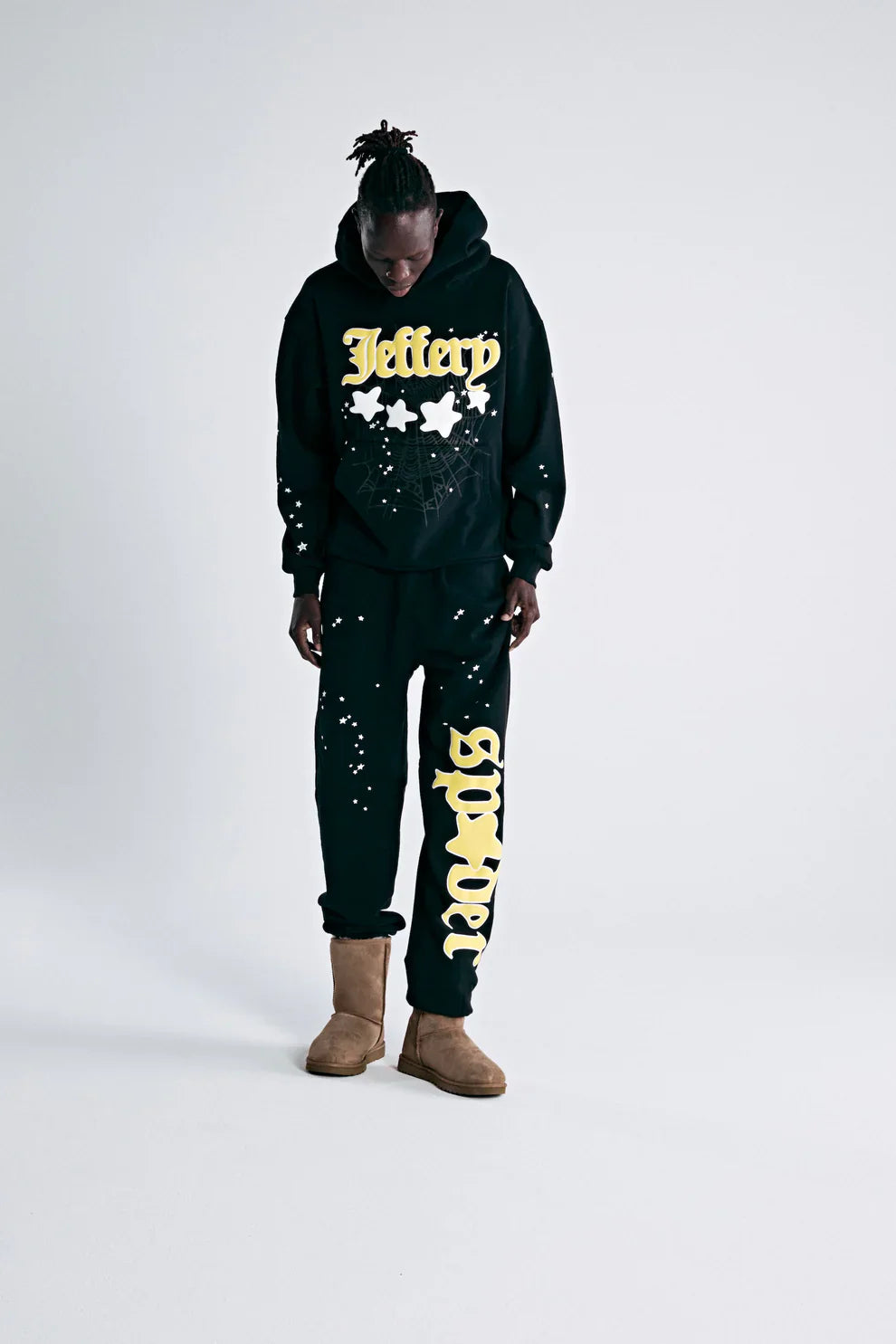 A person in a black hoodie with Jeffery and yellow stars, 100% Cotton Sp5der Sweatpants for Men—Black Streetwear Jogger Pants with Yellow Logo Graphic (relaxed fit), and brown boots stands looking down against a plain white background.