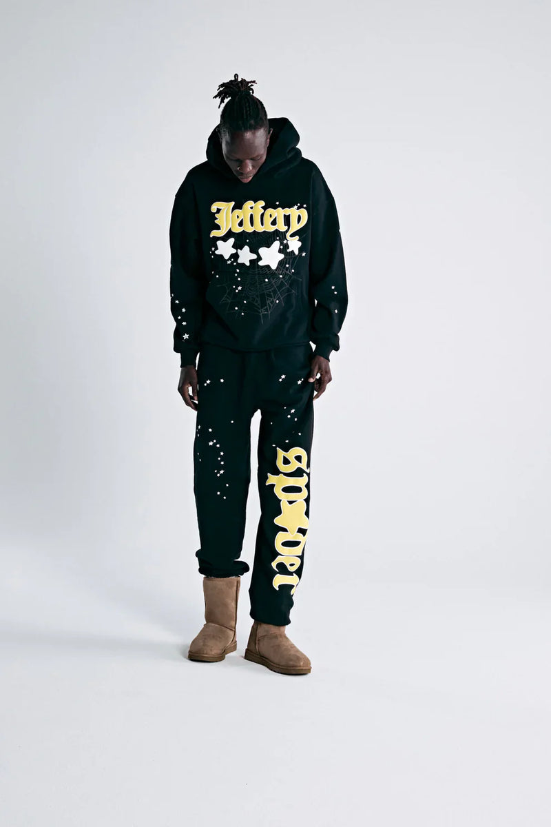 A person in a black hoodie with Jeffery and yellow stars, 100% Cotton Sp5der Sweatpants for Men—Black Streetwear Jogger Pants with Yellow Logo Graphic (relaxed fit), and brown boots stands looking down against a plain white background.