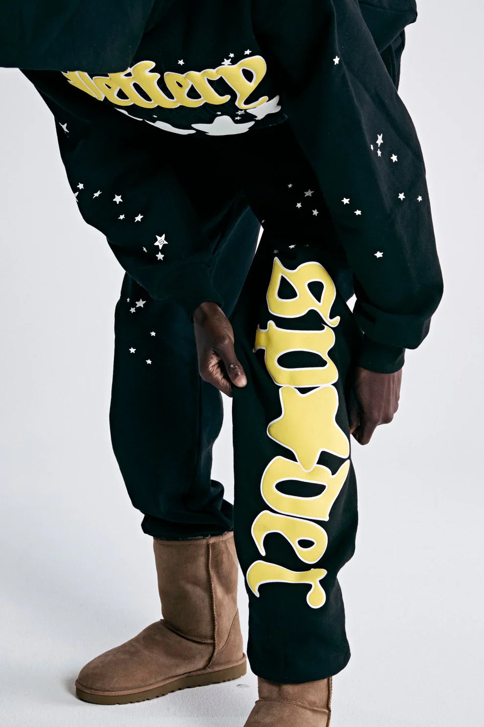 A person wears 100% Cotton Sp5der Sweatpants for Men—black streetwear jogger pants with a yellow logo graphic and relaxed fit—paired with a matching sweatshirt and tan boots, standing out against a plain white background.
