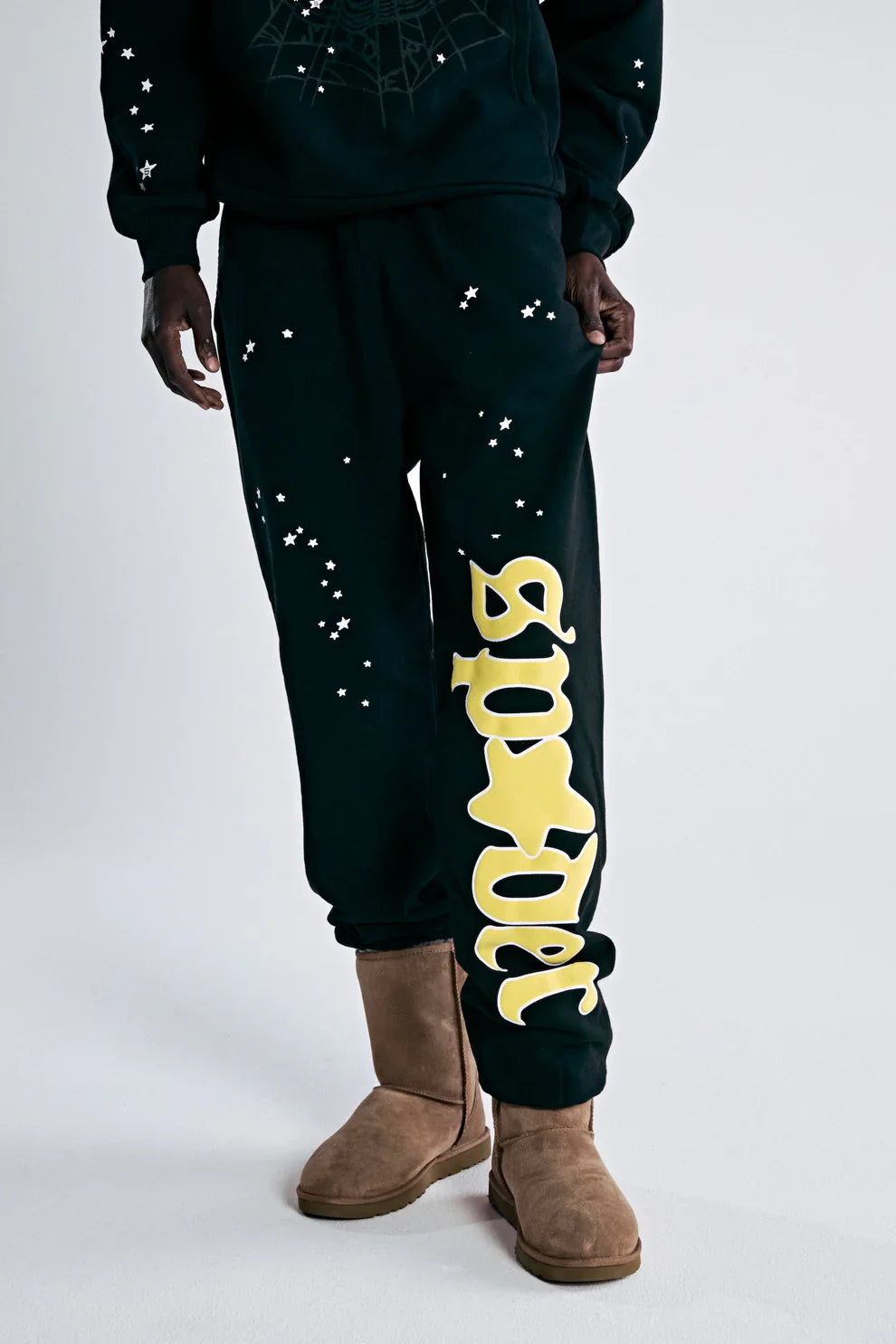 A person wearing 100% Cotton Sp5der Sweatpants for Men—Black Streetwear Jogger Pants with a Yellow Logo Graphic and relaxed fit—paired with a black sweatshirt and tan suede boots for a cool streetwear look.