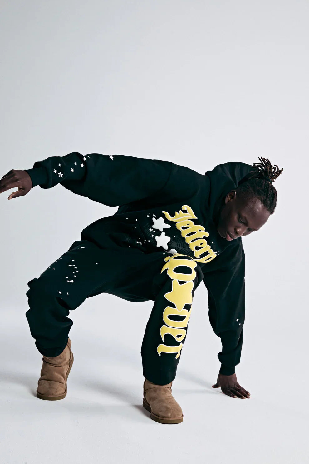 A person in black 100% Cotton Sp5der Sweatpants for Men—streetwear jogger pants with a yellow logo graphic and relaxed fit—poses dynamically. They wear a black hoodie, tan boots, have short twists, and stand against a white background.