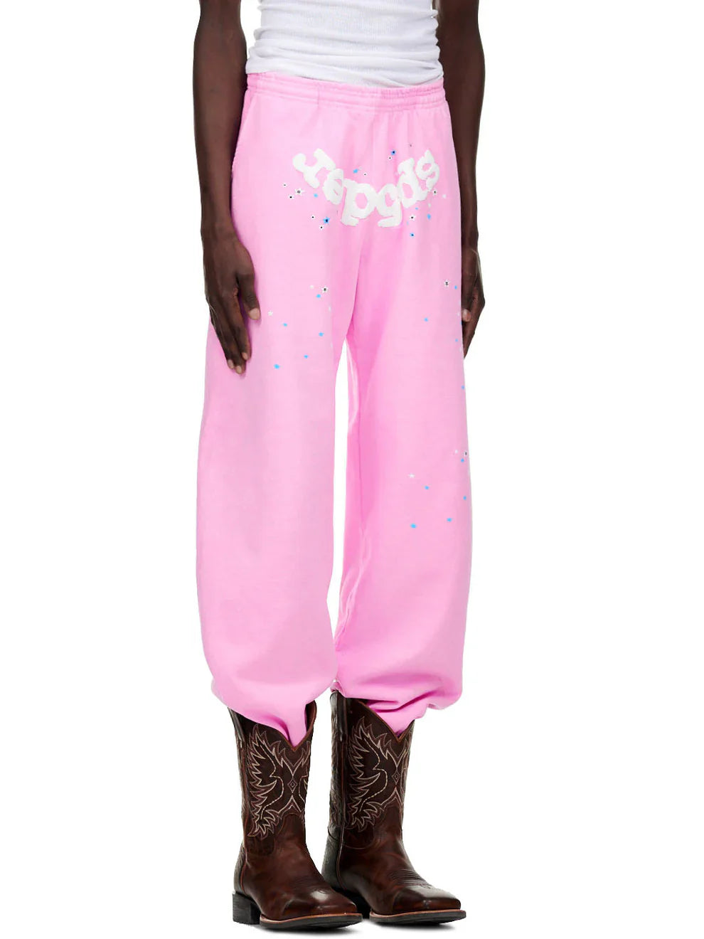 A person wears Sp5der Sweatpants – unisex light pink cotton sweatpants with a front logo and star print, paired with a white sleeveless shirt and dark brown cowboy boots. Only the body from shoulders down is visible.