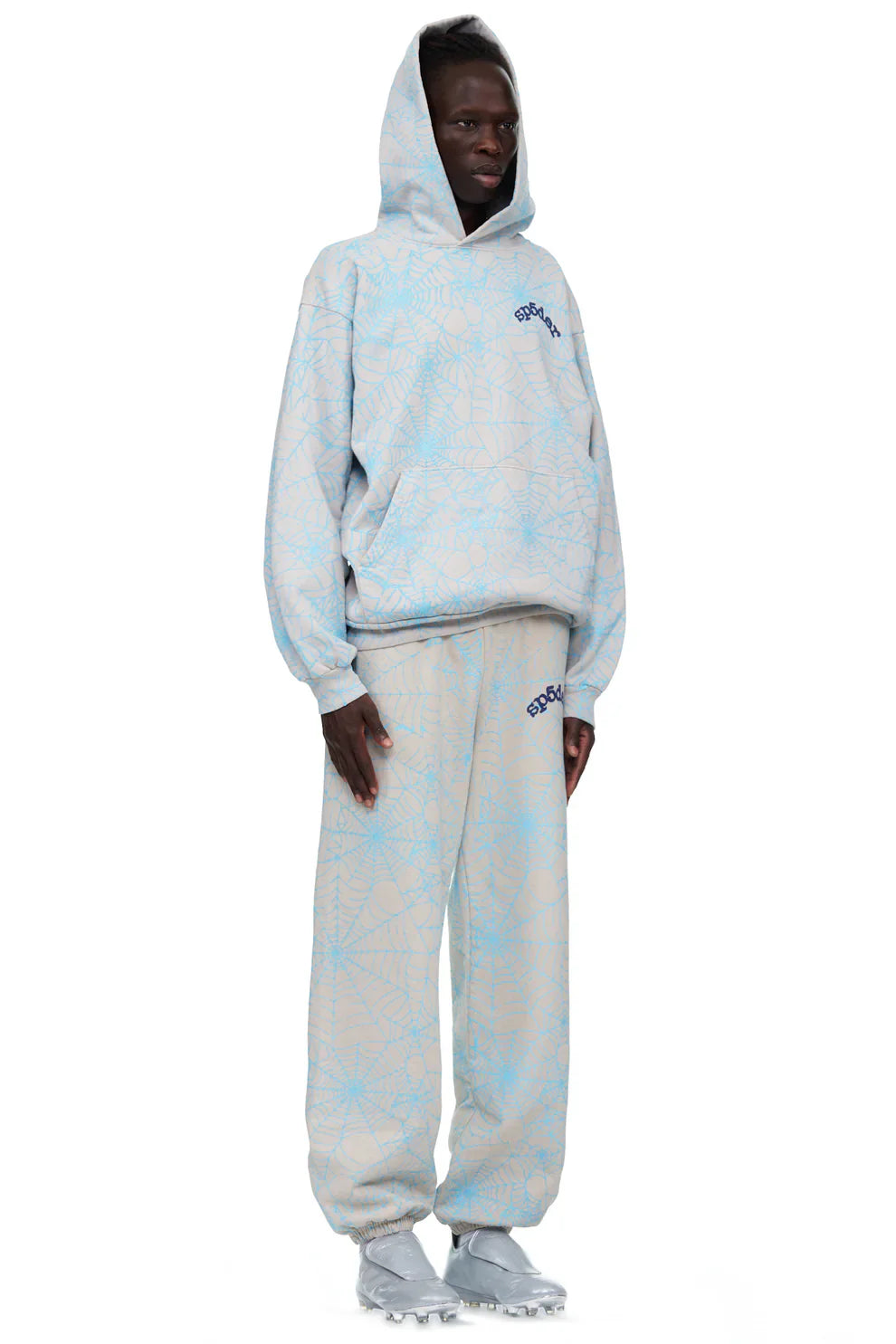 A person stands wearing light grey 100% cotton Sp5der sweatpants for men with an all-over spider web print and a relaxed fit.