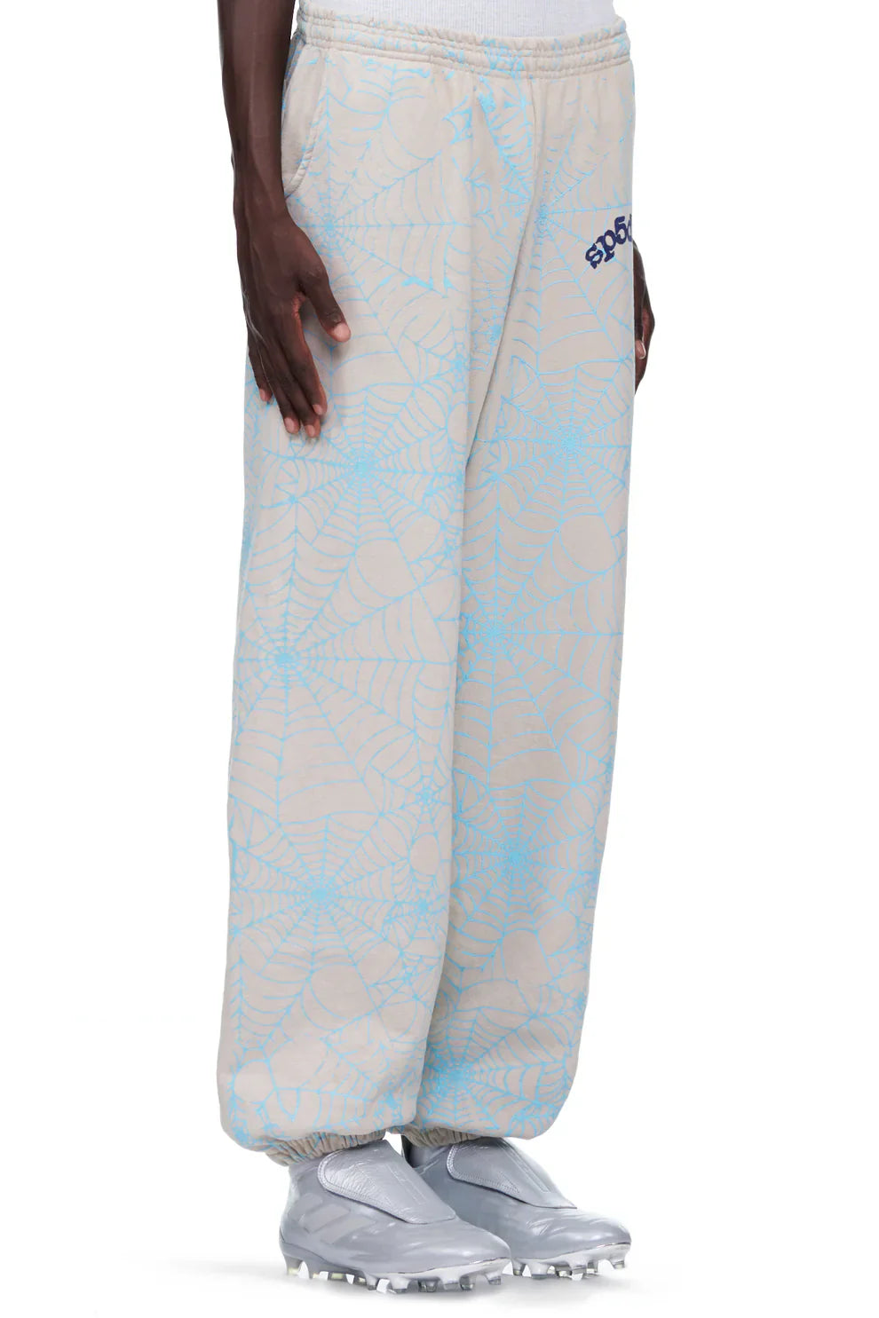 A person wears 100% Cotton Sp5der Sweatpants for Men—light grey jogger pants with an all-over blue spider web print, relaxed fit, and visible SP5DER logo on the left hip. The streetwear sweatpants are highlighted; upper body not shown.