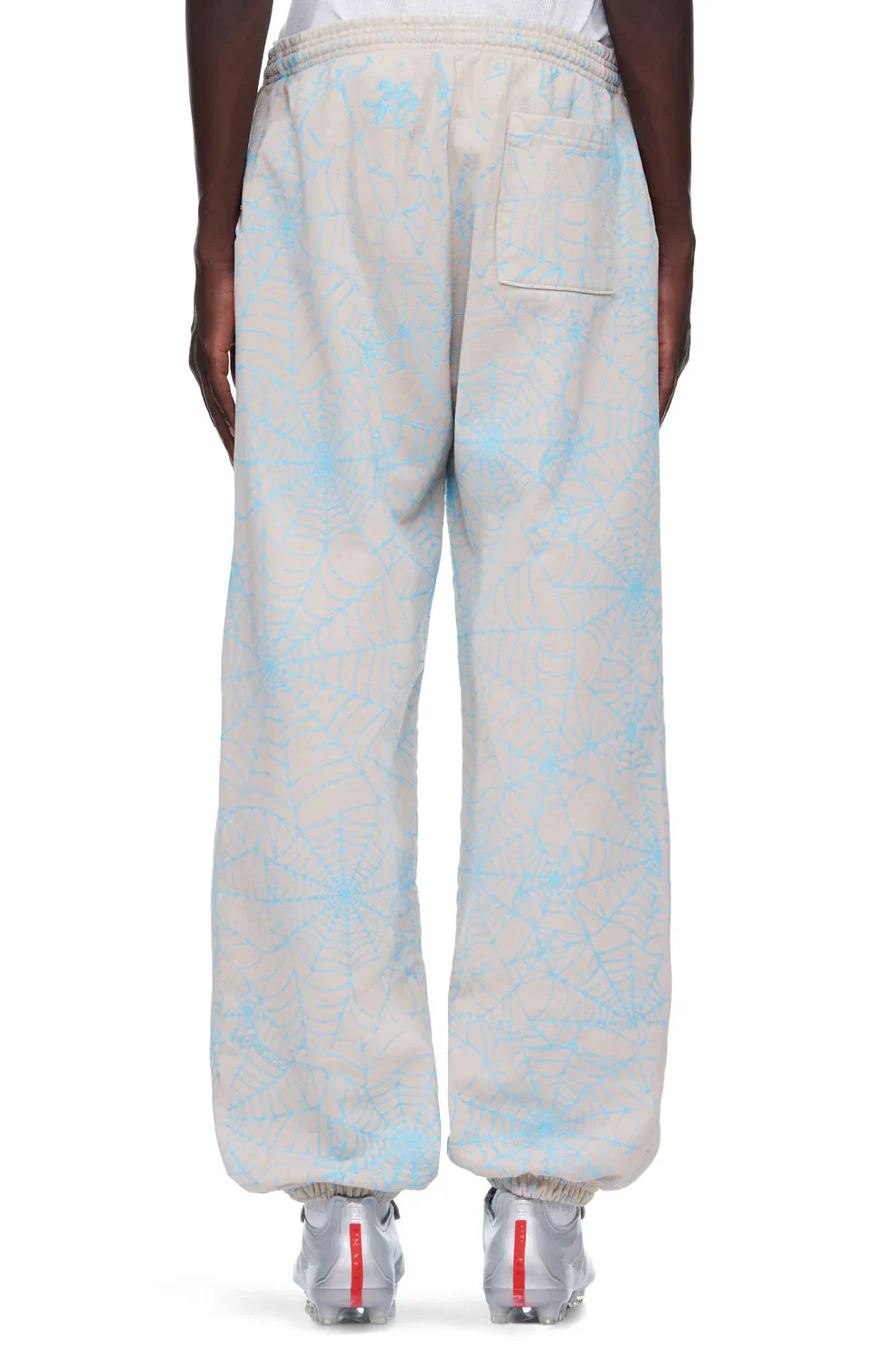 A person wears 100% Cotton Sp5der Sweatpants for Men in light grey with an all-over spider web print and relaxed fit, featuring an elastic waistband, back pocket, and ankle cuffs. White sneakers complete this streetwear look.