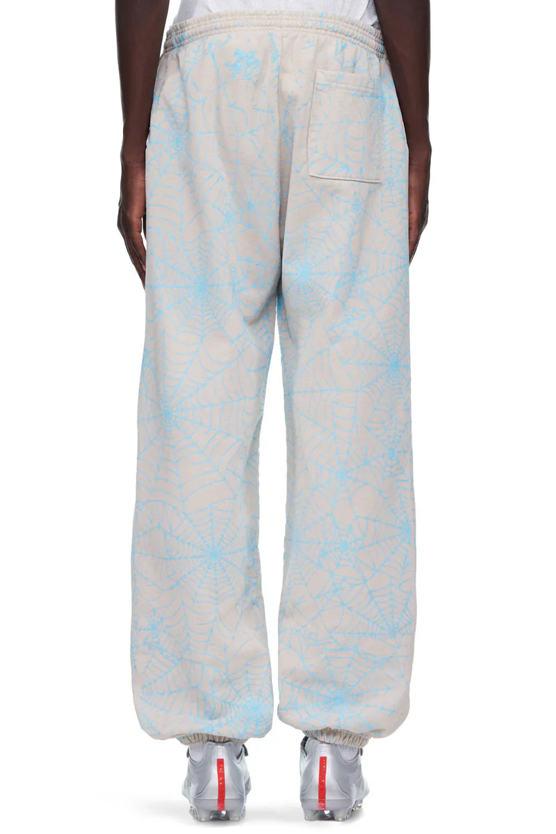 A person wears 100% Cotton Sp5der Sweatpants for Men in light grey with an all-over spider web print and relaxed fit, featuring an elastic waistband, back pocket, and ankle cuffs. White sneakers complete this streetwear look.