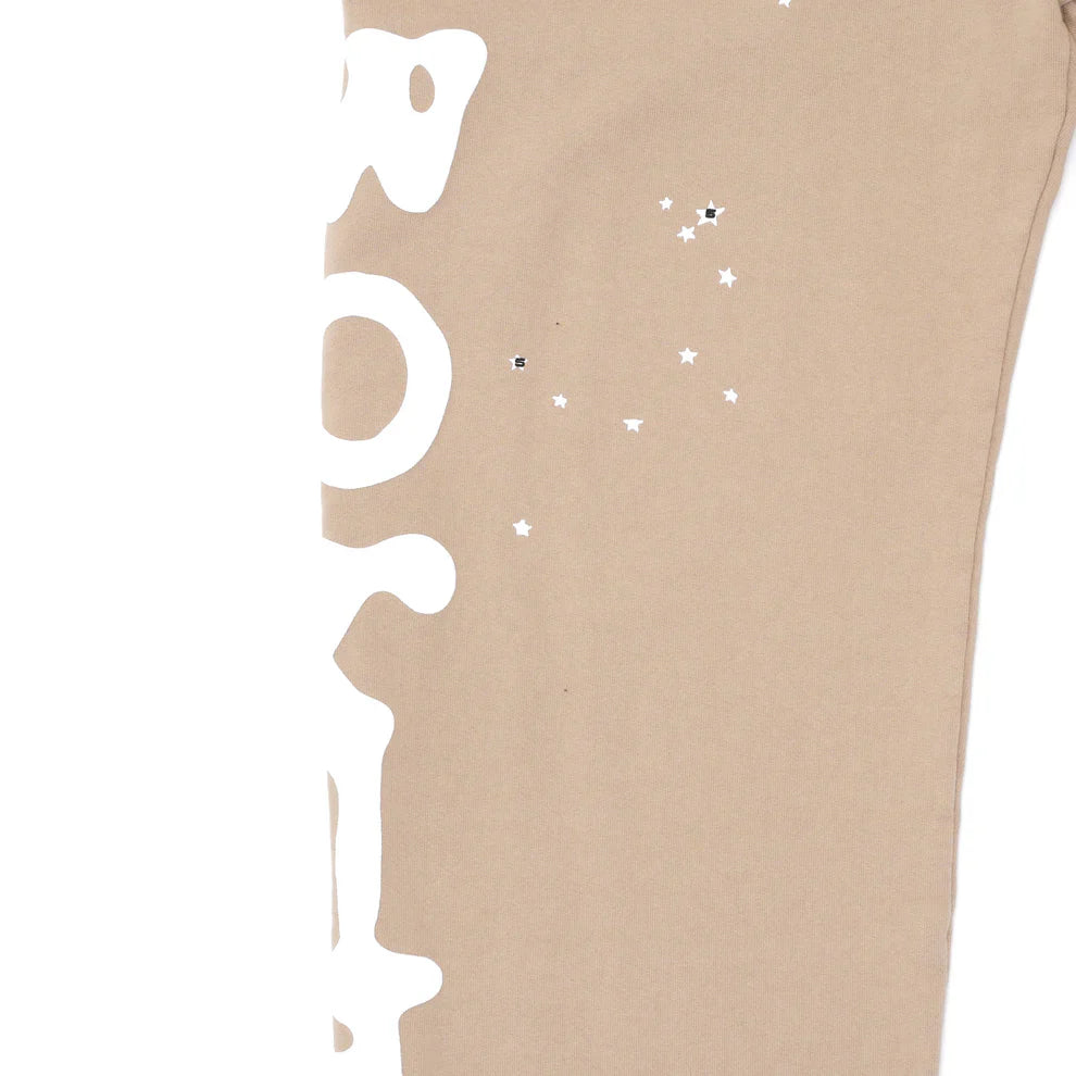 A close-up of the 100% Cotton Sp5der Sweatpants for Men in beige shows large, white, partially visible side logo graphics on the left and a cluster of small white and black star shapes on the right.